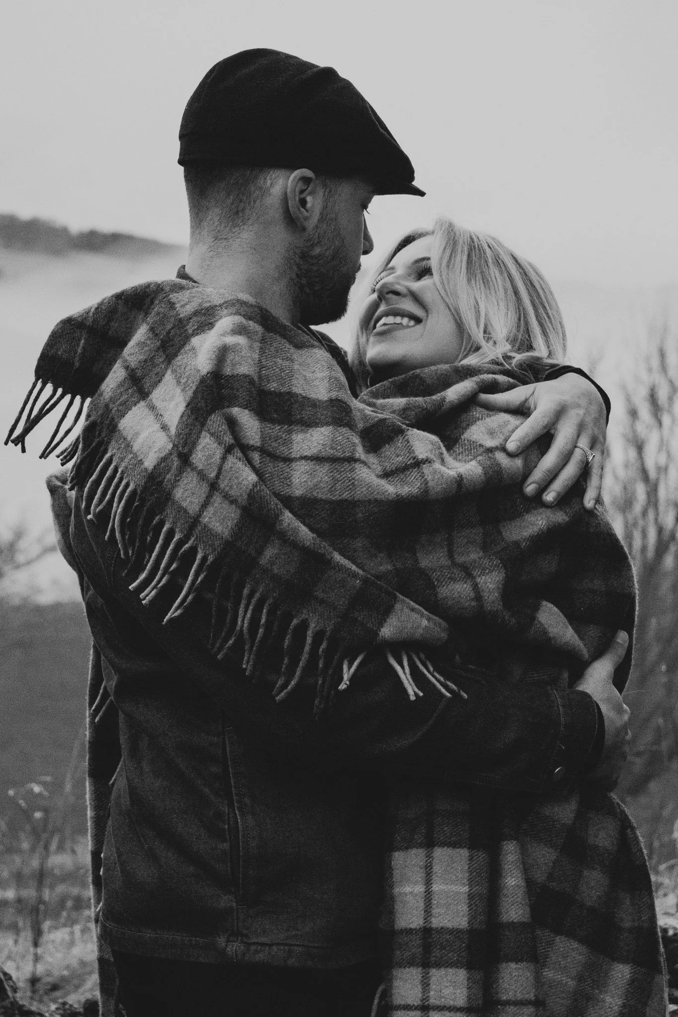 A high-contrast black and white photograph of a couple hugging tightly, with the man wearing a traditional flat cap wrapped in a traditional Scottish tartan blanket.