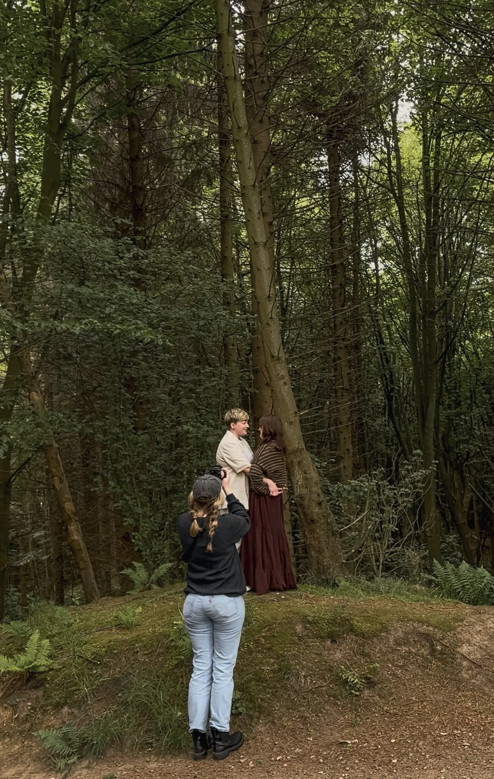 A wide landscape portrait of a photographer capturing the intimate embrace between a couple standing beautifully along a hidden path deep inside a lush green Scottish woodland.