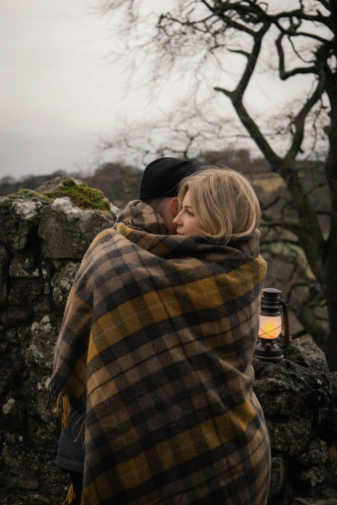 An intimate couple wrapped together in a traditional Scottish tartan blanket, holding a warm glowing lantern outdoors at dusk.