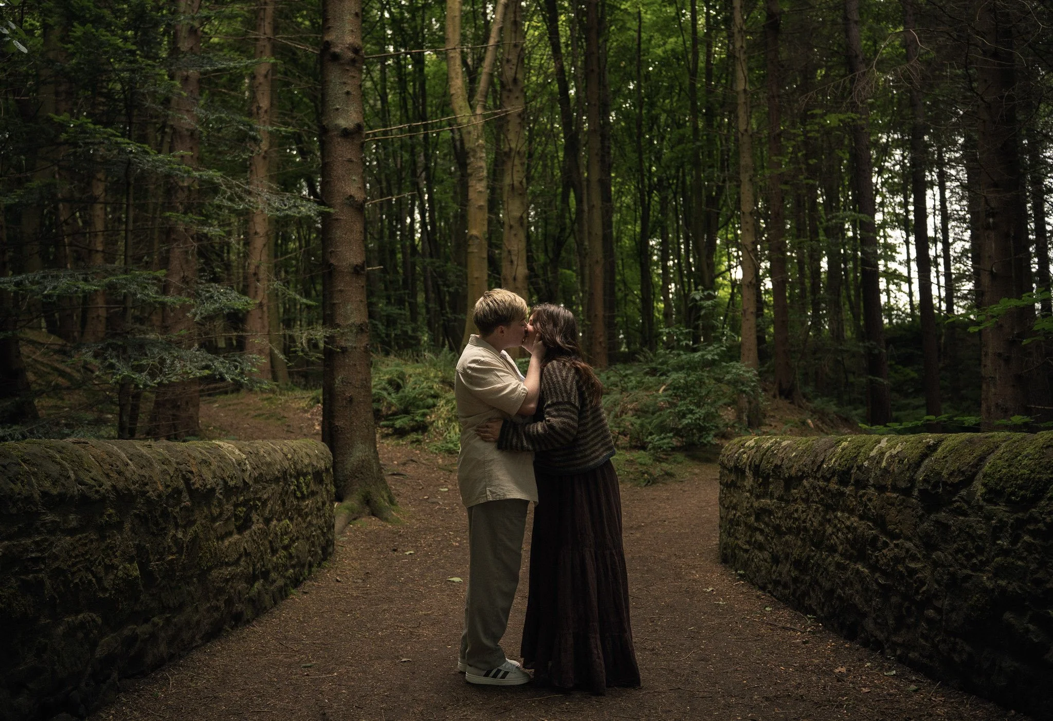 A couple embracing closely surrounded by tall, dark pine trees in an enchanted Scottish forest.