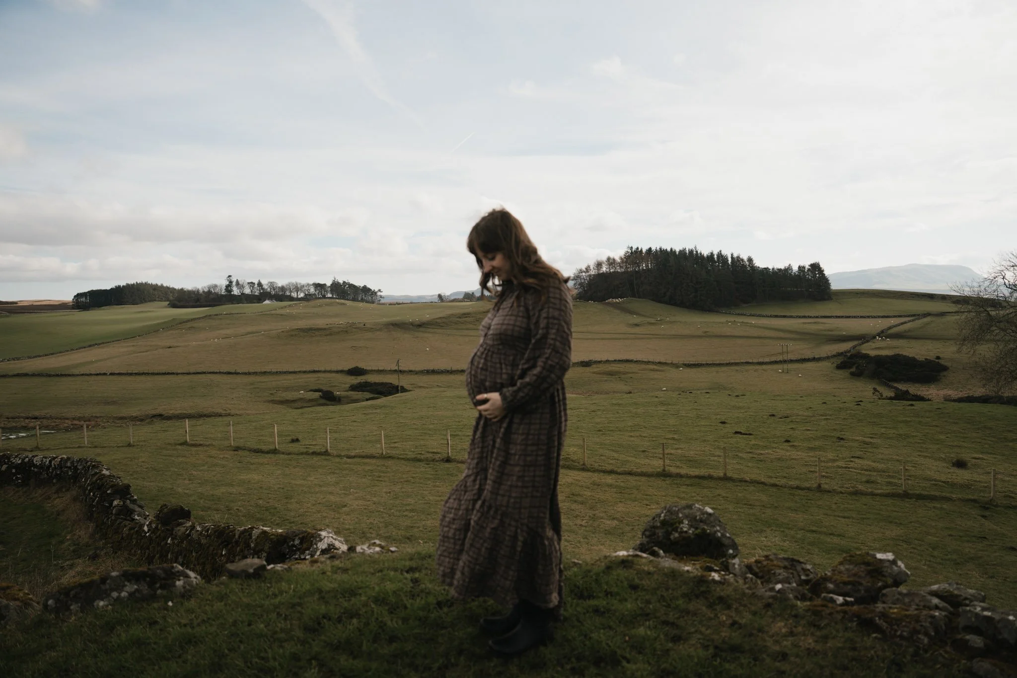 A lone pregnant woman in a flowing dress standing on a vast, open moorland plain in Scotland beneath a moody dramatic sky.