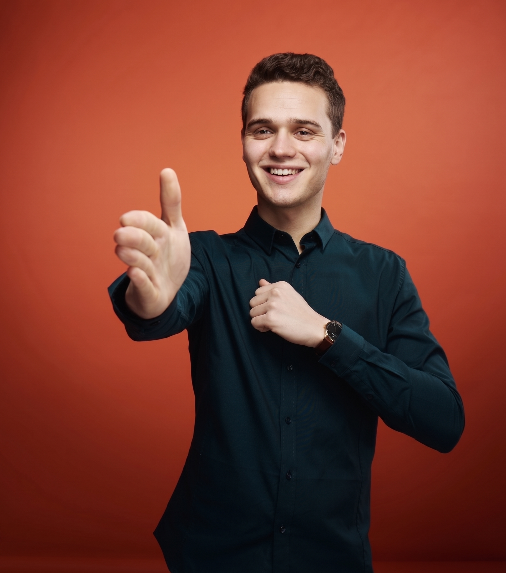 Ein junger Mann in dunklem Hemd mit einer Uhr am Handgelenk zeigt mit einem freundlichen Lächeln auf den Betrachter, während er eine Hand in die Kamera hält, gegen einen orangefarbenen Hintergrund.