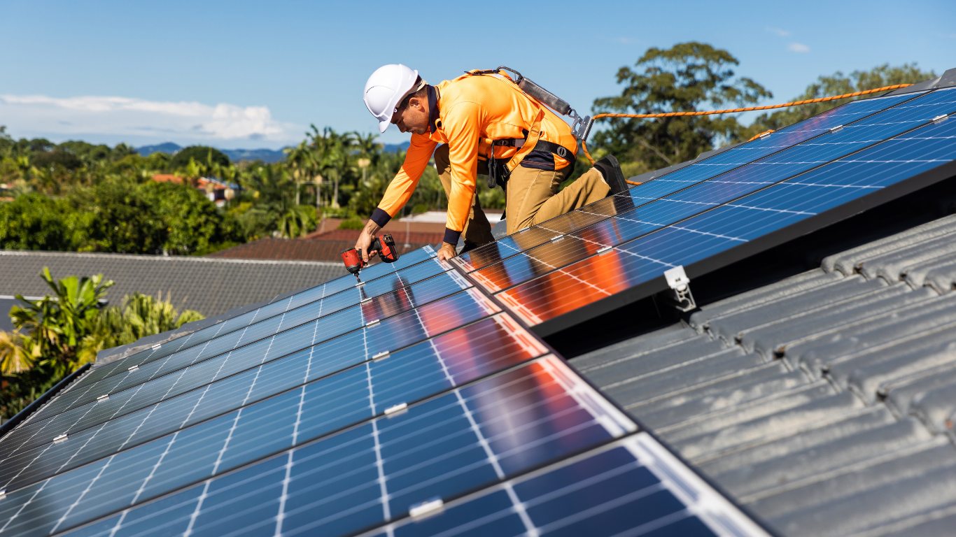 Un técnico trabaja en la instalación de paneles solares en el techo de una casa, usando equipo de protección y herramientas.