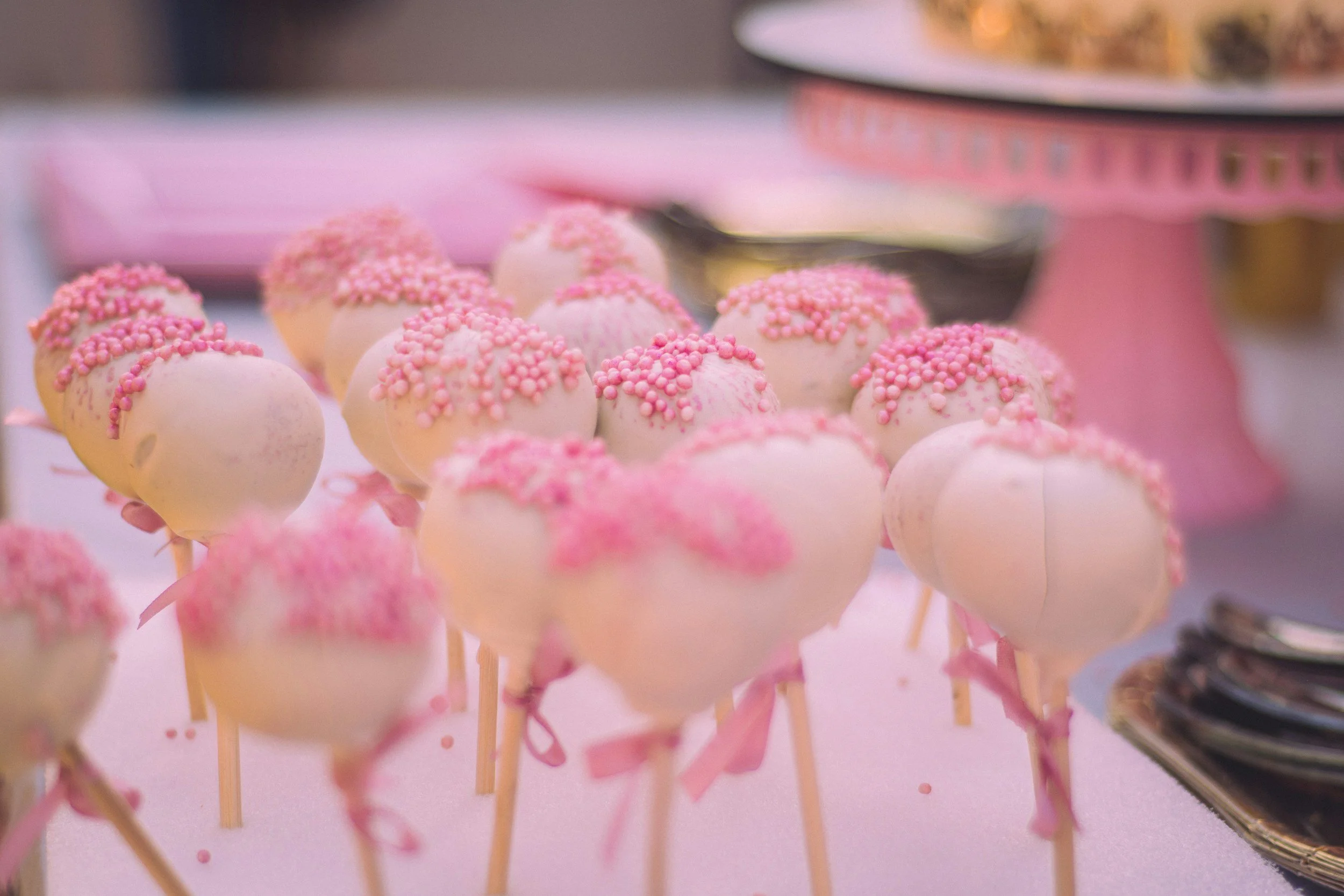 Pink cake pops decorated with pink sprinkles on wooden sticks, arranged on a pink surface with a cake stand in the background.