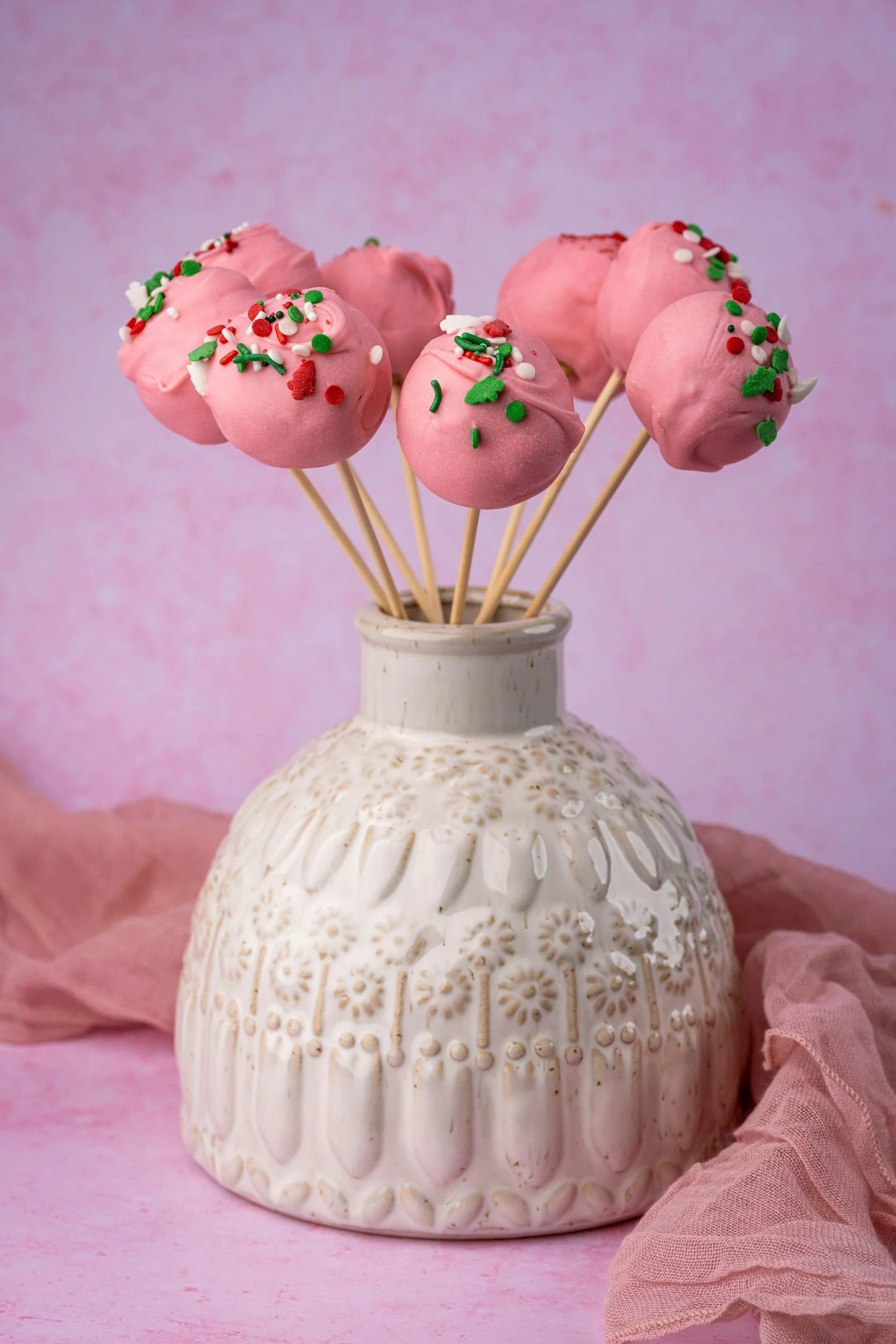 Pink cake pops decorated with colorful sprinkles and placed in a white ceramic vase, with a pink fabric underneath and a pink and purple background.