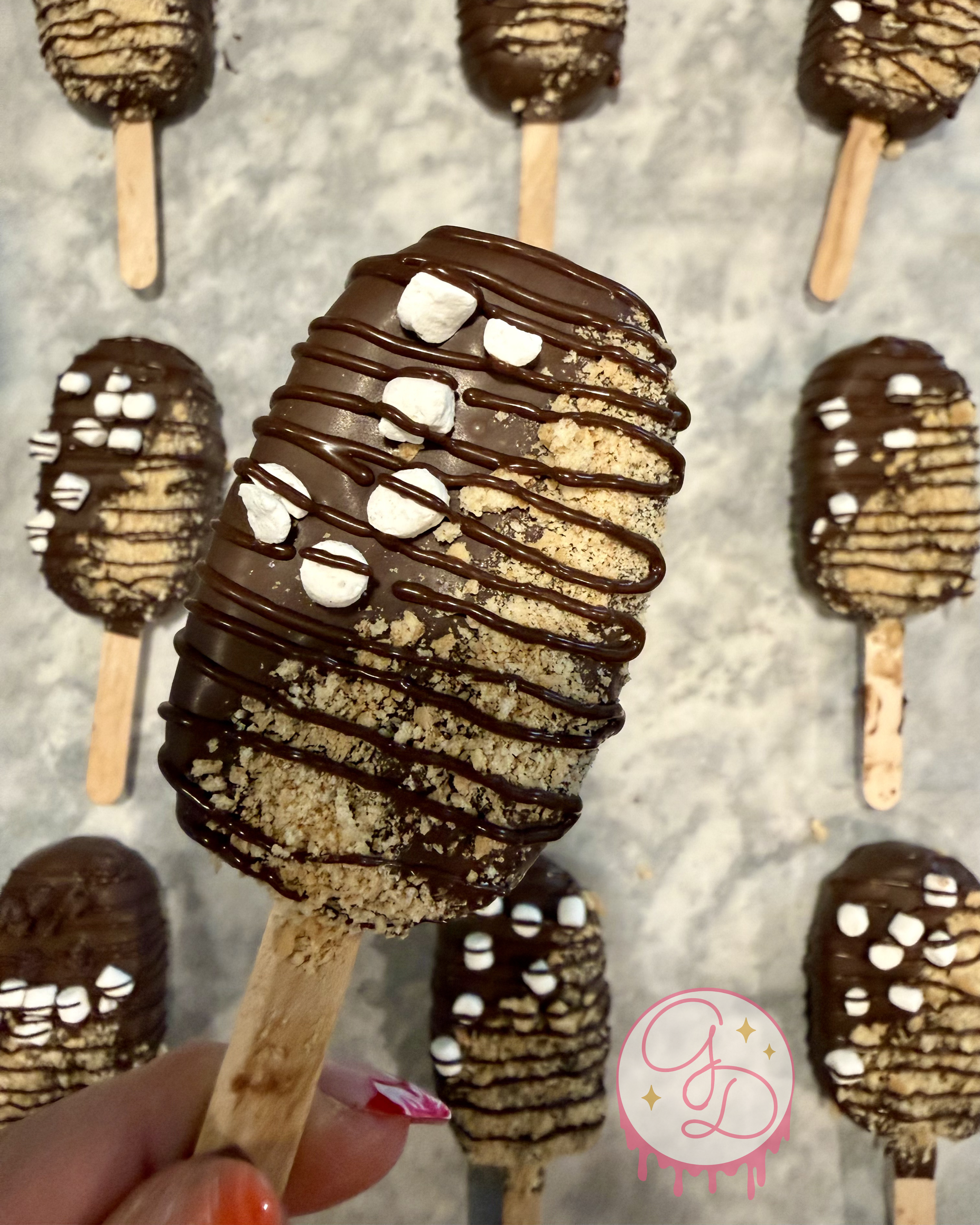 Chocolate-covered ice cream bars on wooden sticks, topped with mini marshmallows and drizzled with chocolate. Several ice cream bars arranged on a sheet, some with sprinkled toppings.