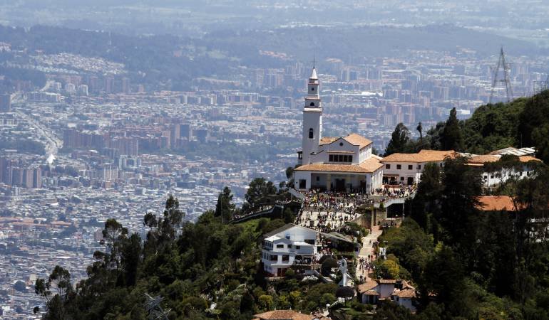Vista panorámica de una iglesia en una colina rodeada de árboles, con una ciudad extensa en el fondo.