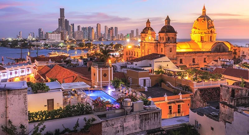 Vista urbana al atardecer con edificios modernos y una iglesia colonial con cúpulas en primer plano, cerca de un río con barcos, en una ciudad costera