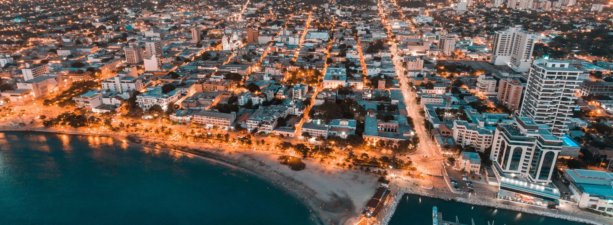 Vista aérea de una ciudad costera en la noche, con edificios altos y calles iluminadas, incluyendo una playa con palmeras en primer plano.