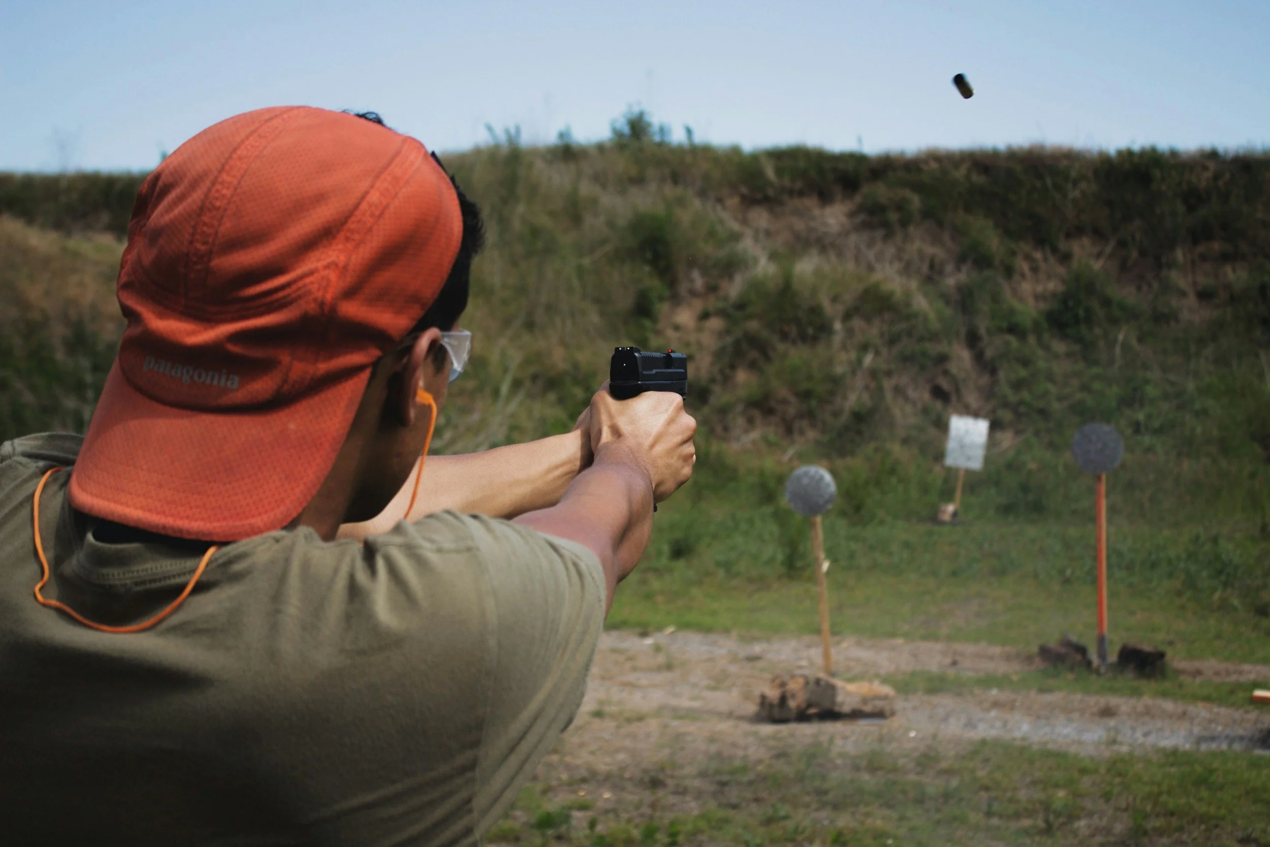 Person wearing orange hat and safety glasses aiming a handgun at a shooting range with targets in the background.