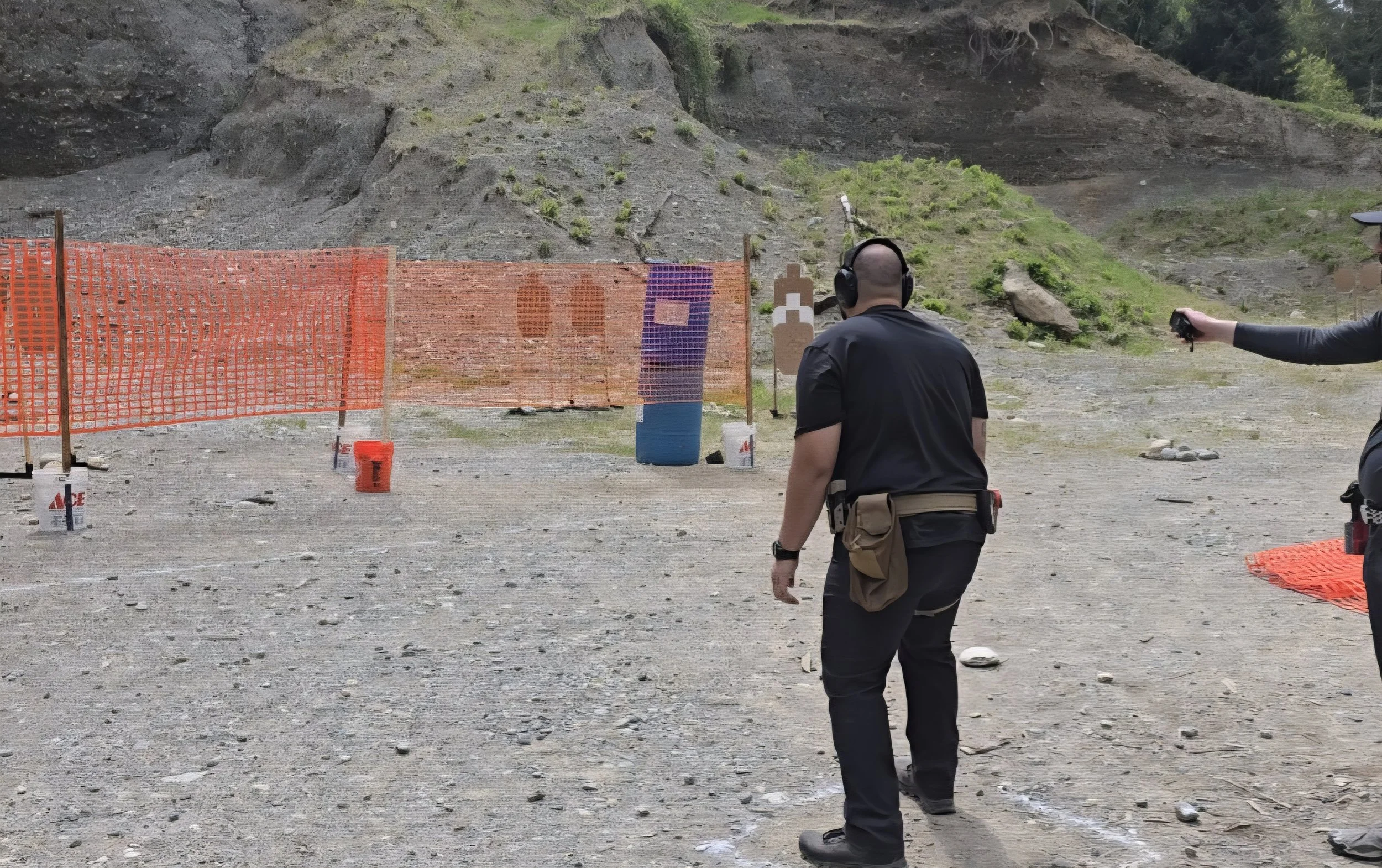 Two people at a shooting range outdoors with targets, orange safety fencing, and a mountainous background, one man facing away with hearing protection and another person holding a firearm.