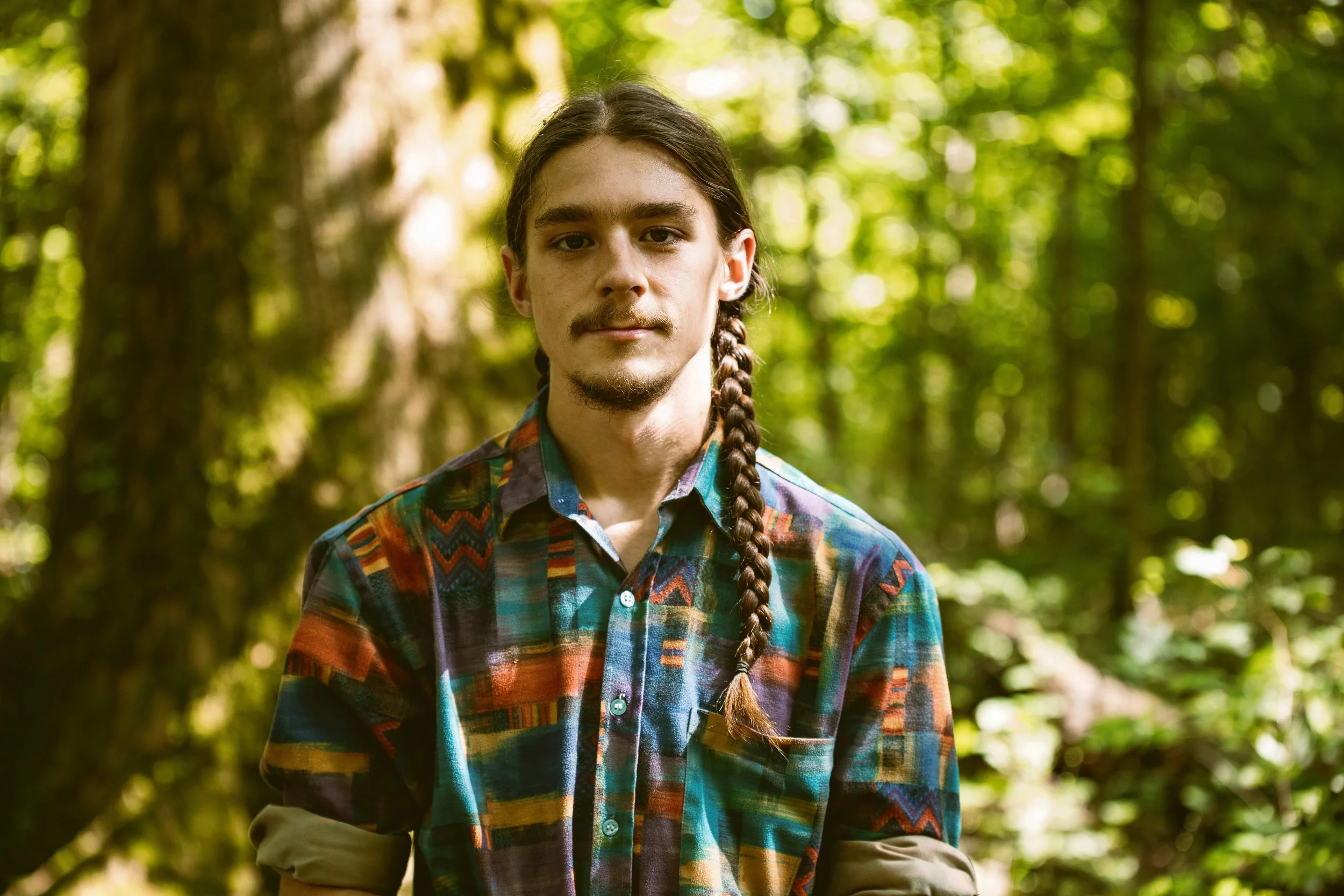 A young man with long hair in a braid, wearing a colorful plaid shirt, standing in a green forest.