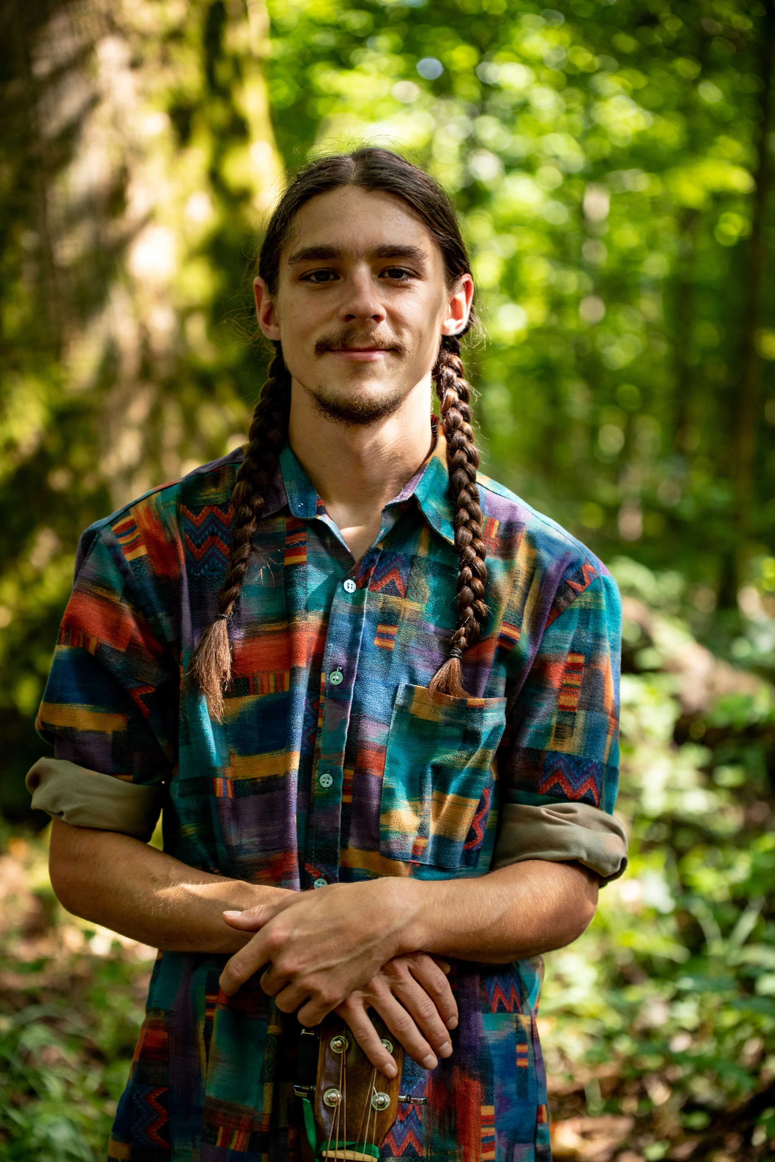A young man with long brown hair in two braids, wearing a colorful patterned shirt with rolled-up sleeves, standing outdoors in a lush green forest. He has a subtle smile and is holding a ukulele with his left hand while crossing his arms.