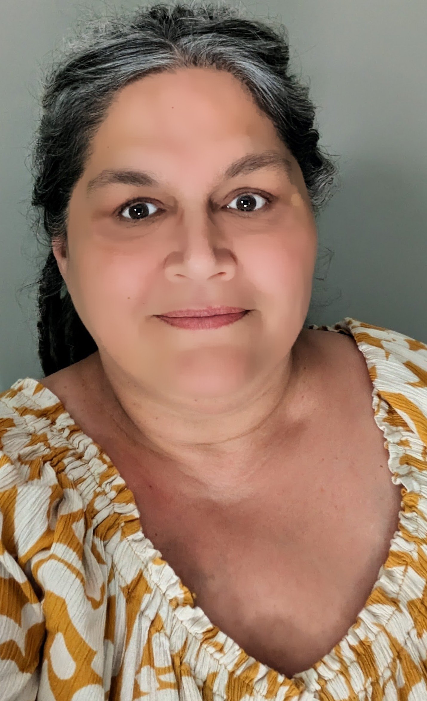 Close-up of a middle-aged woman with dark gray hair, smiling, wearing a yellow and white patterned blouse, against a plain gray background.