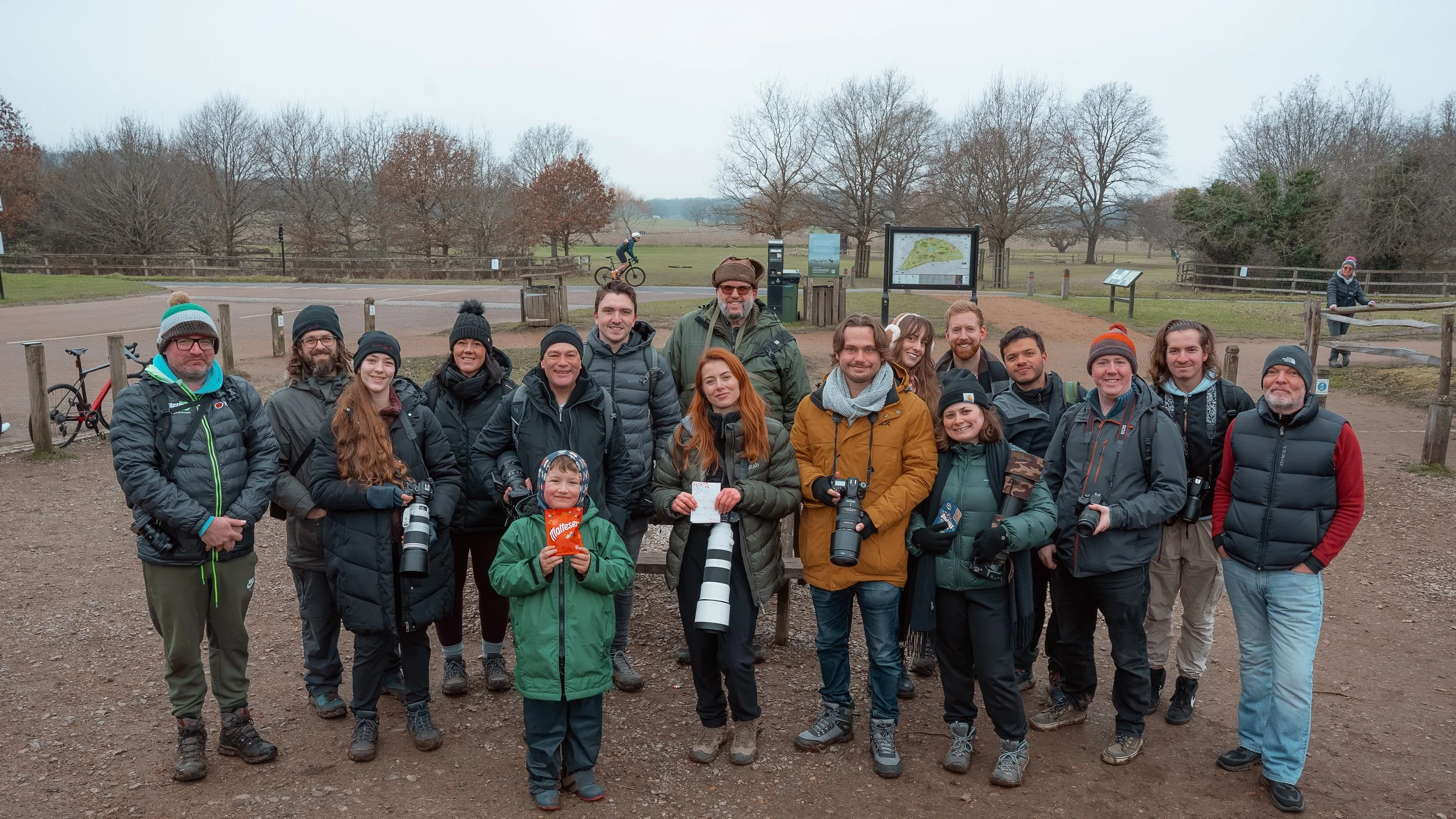 Group of people in outdoor gear posing for a photo in a park or nature reserve, with trees and informational signs in the background.