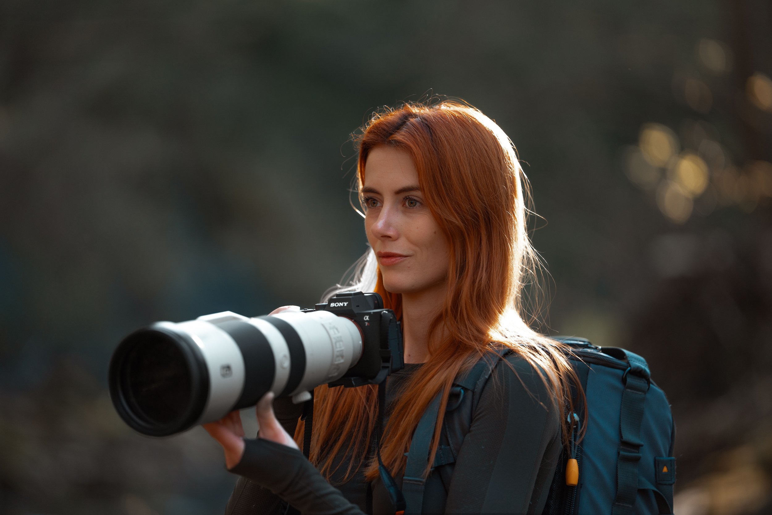 A woman with red hair holding a large telephoto camera lens, wearing a blue backpack, in an outdoor setting with blurred dark background.
