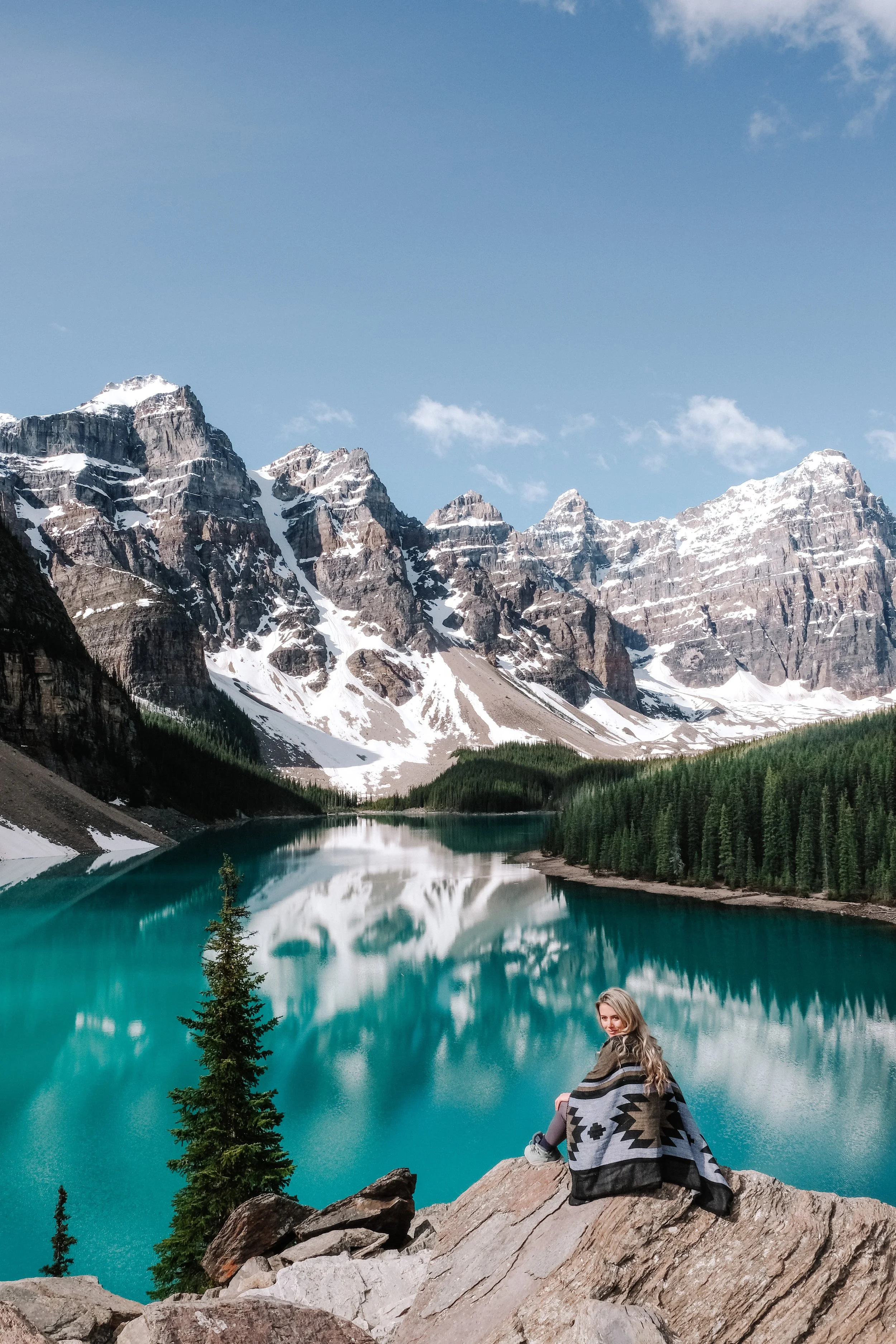 A woman sitting on rocks by a turquoise lake, surrounded by pine trees and snow-capped mountains under a blue sky with clouds.