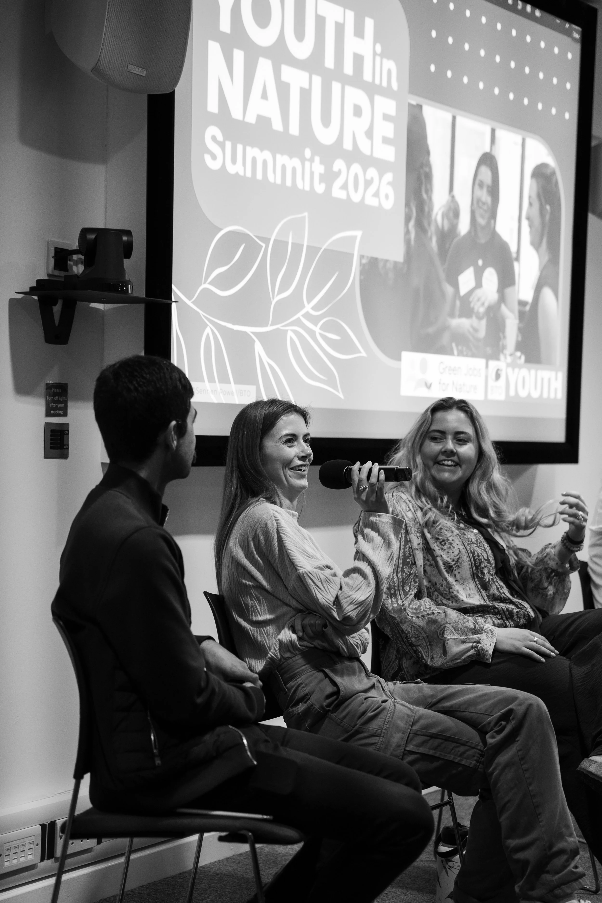 Four people are sitting on chairs in a panel discussion, with three women engaged in conversation, one holding a microphone. There is a projection screen behind them displaying a logo and text for the Youth in Nature Summit 2026, with a logo depicting leaves.