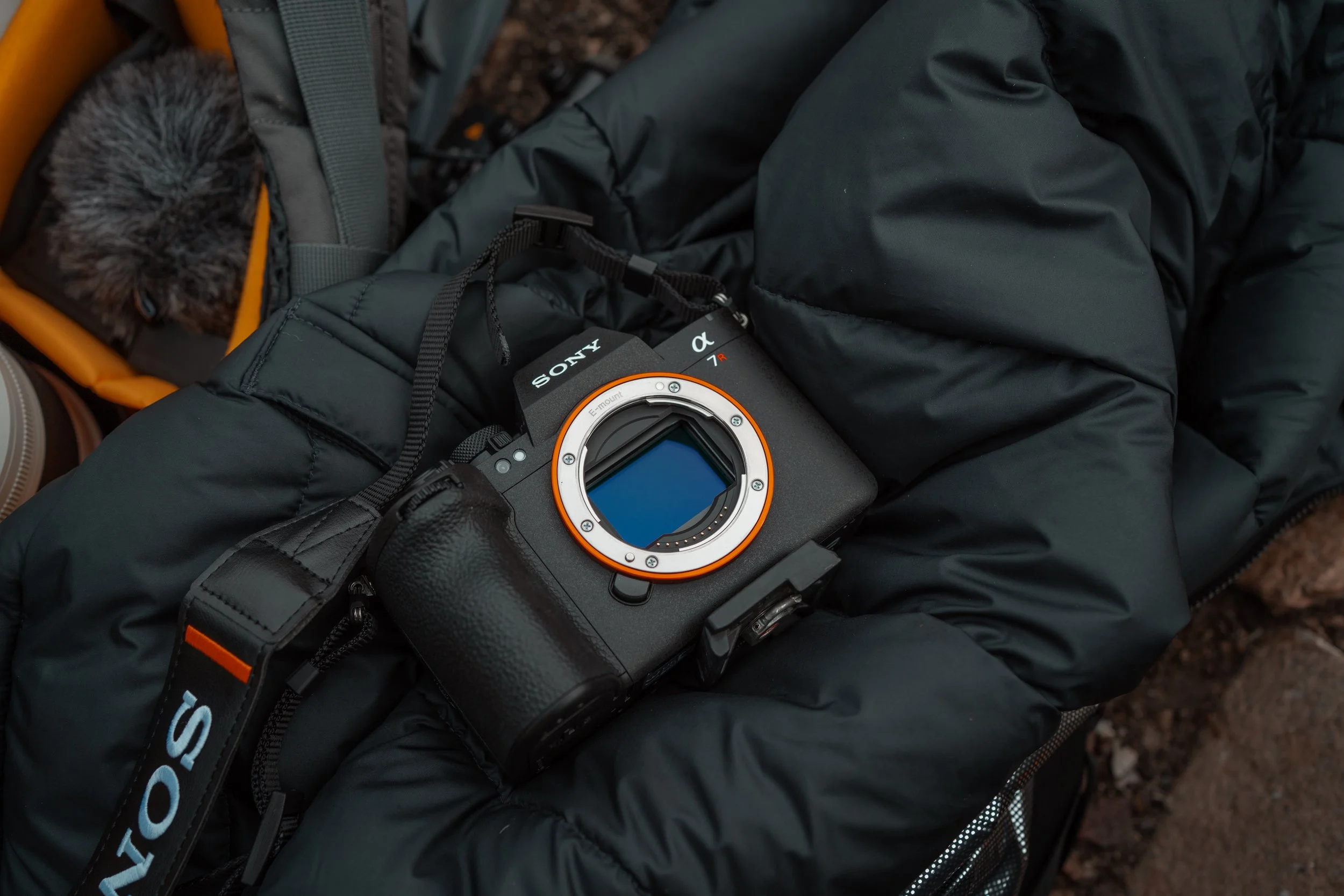 A Sony mirrorless camera with a detachable lens placed on a black jacket, next to a grey backpack with a fur-lined hooded garment partially visible in the background.