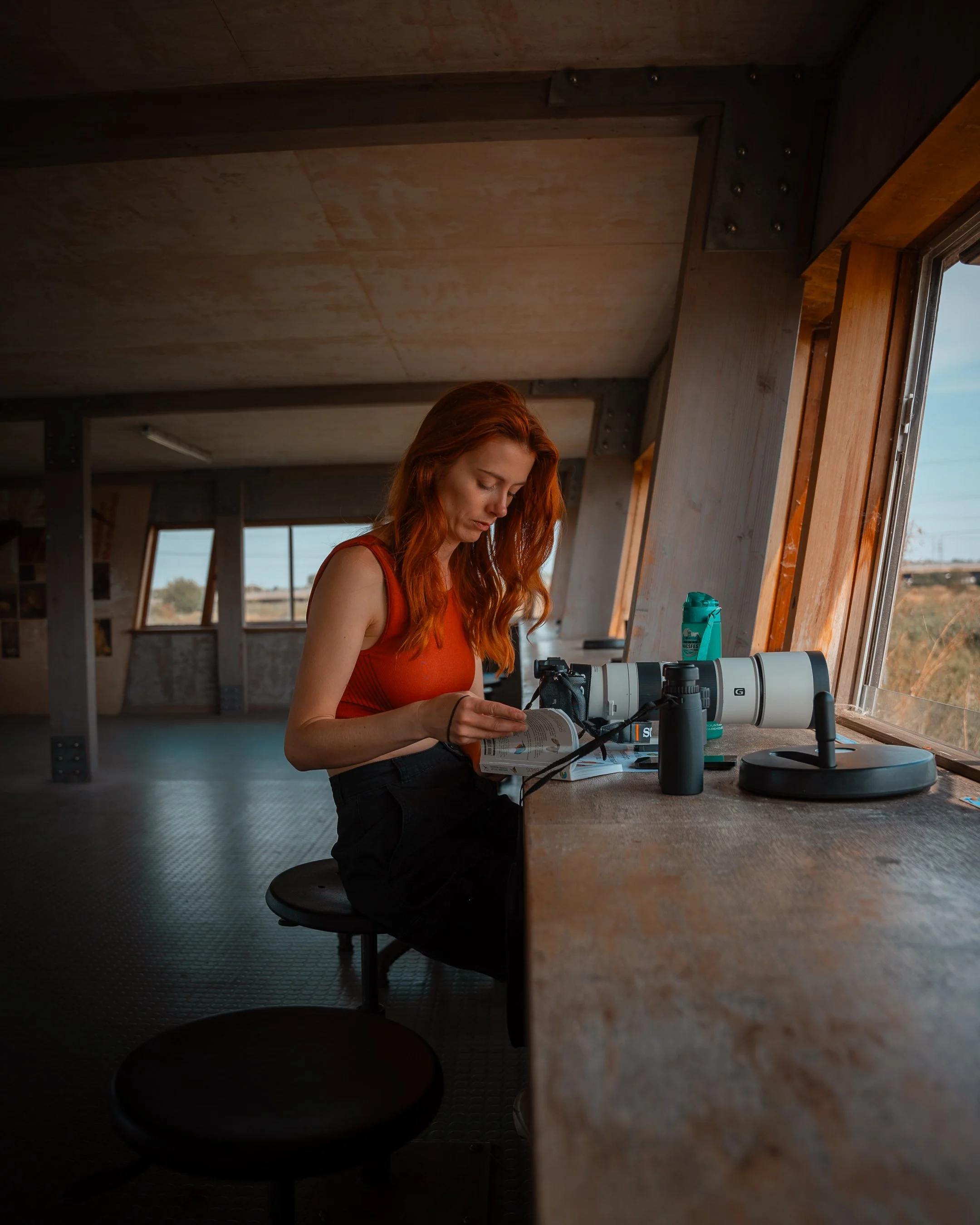 A woman with red hair wearing an orange tank top is sitting at a wooden counter, looking at and reading a booklet. In front of her are a camera, a water bottle, and other equipment. The room has large windows, wooden beams, and a ceiling with a wooden finish, with a rural landscape visible outside.