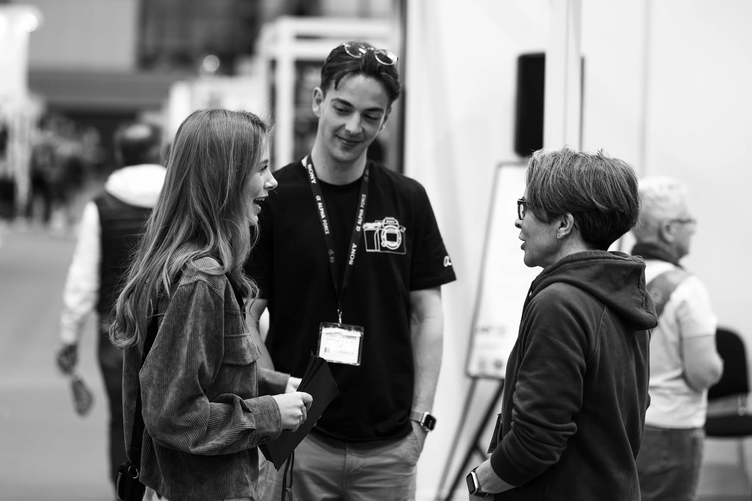 Three people having a conversation at an indoor event. One woman with long hair, another woman with short hair and glasses, and a young man with a lanyard around his neck. The background shows other attendees and a blurred setting.
