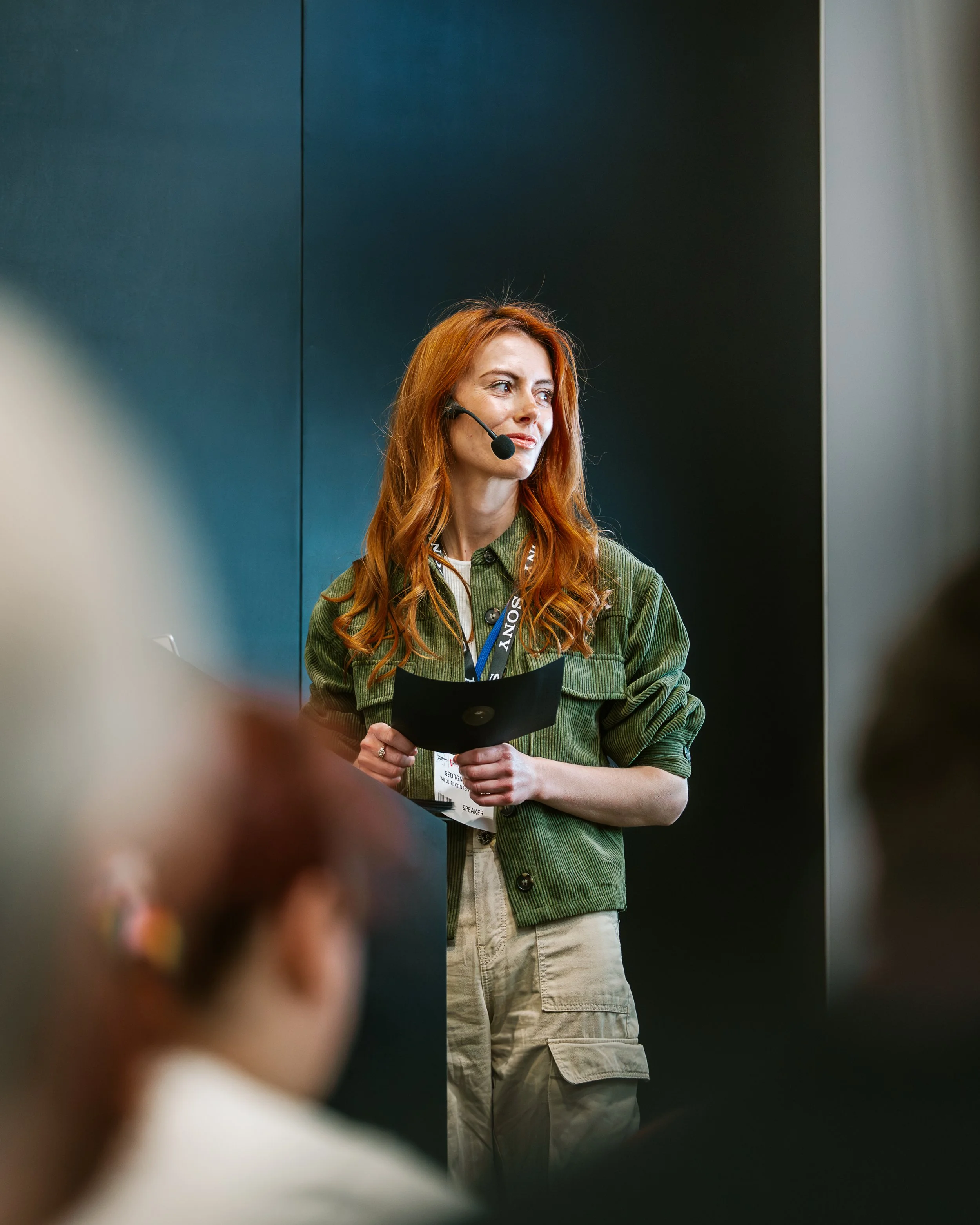 A woman with red hair wearing a green jacket and beige cargo pants standing in front of a dark background, holding a black folder, with a headset on her head, and a name tag hanging around her neck.