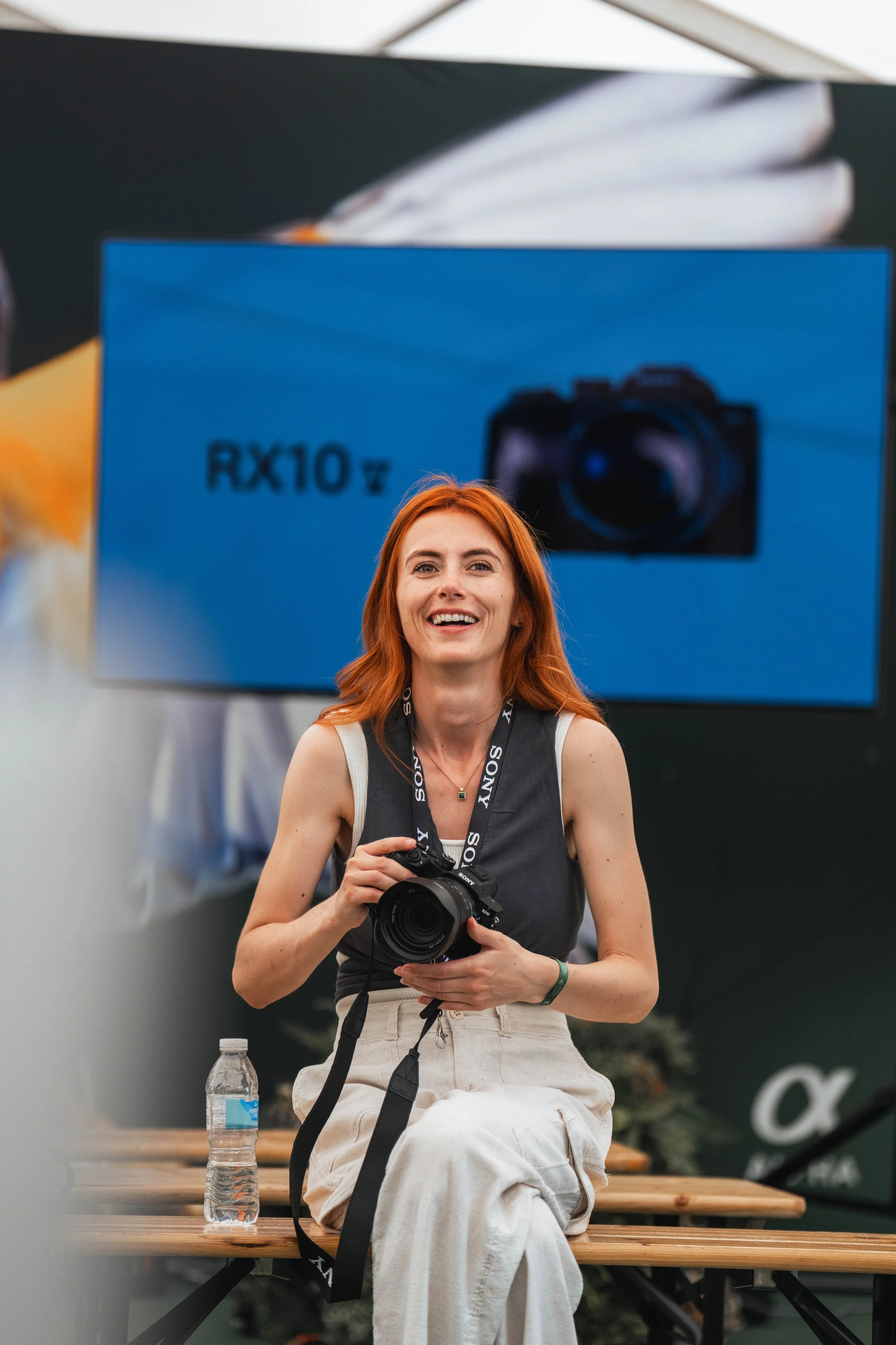 A woman with red hair smiling, holding a camera, sitting on a wooden bench, with a Sony Alpha camera strap around her neck, and a water bottle on the bench beside her. She is at an event with a Sony banner in the background.
