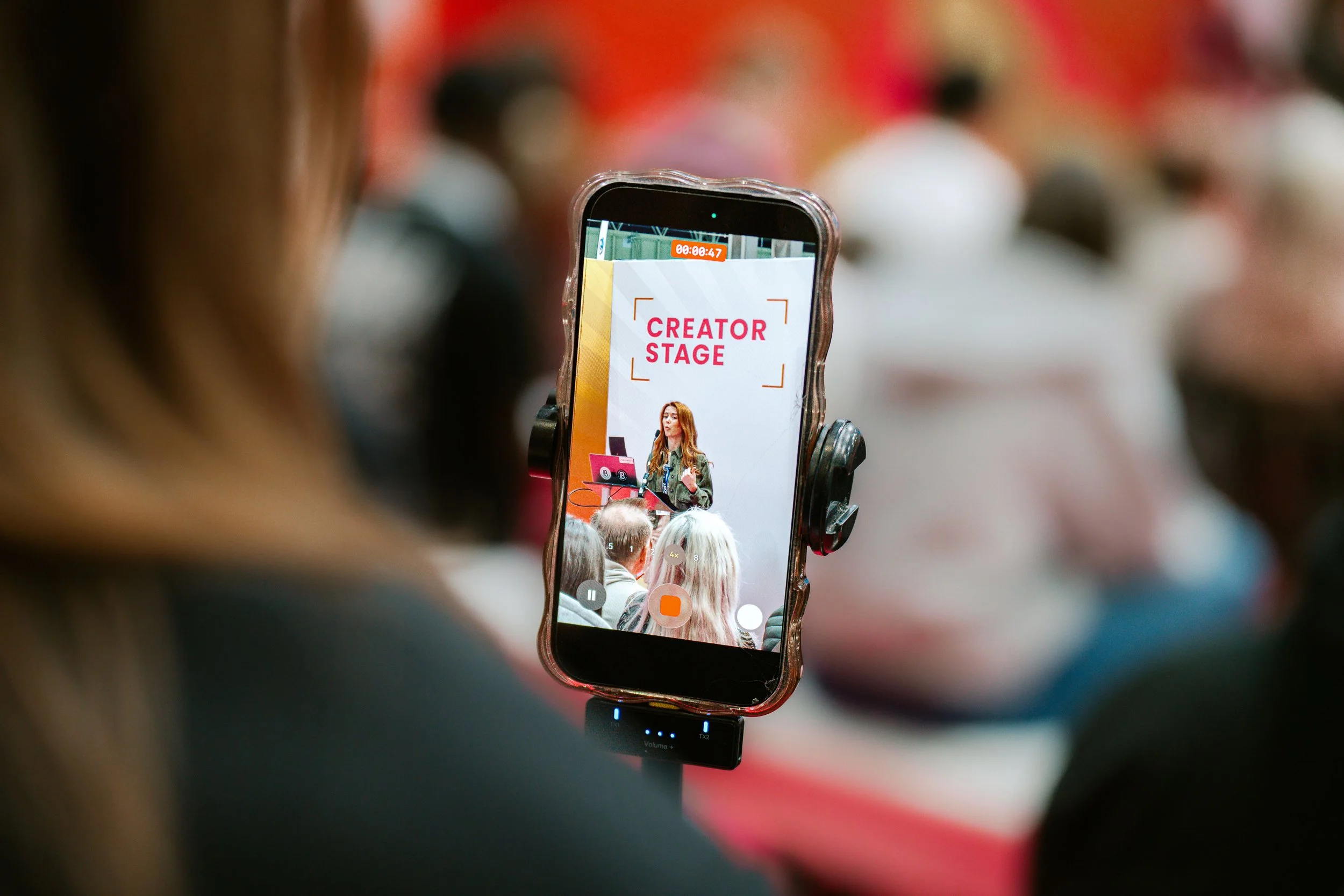 A smartphone camera shows a woman speaking at a stage labeled 'Creator Stage' as seen through a crowd at an event.