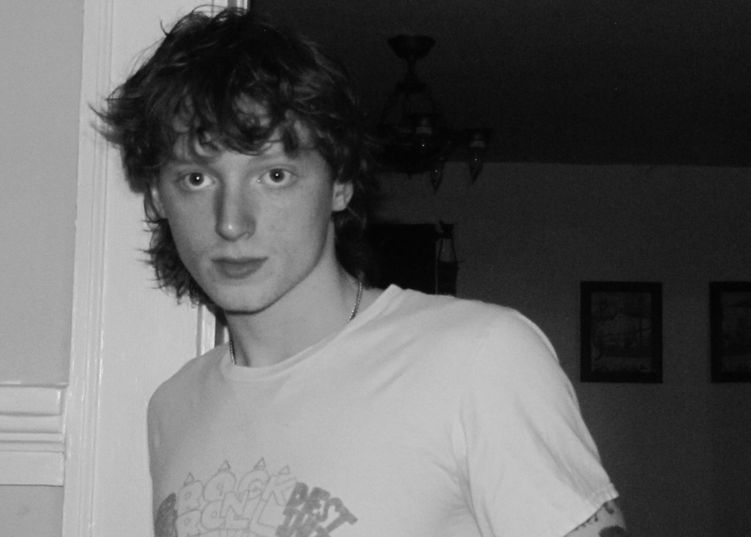 A black-and-white photograph of a young person with curly hair, looking directly at the camera, wearing a white T-shirt with a design on it, standing indoors near a wall with a ceiling light fixture visible in the background.