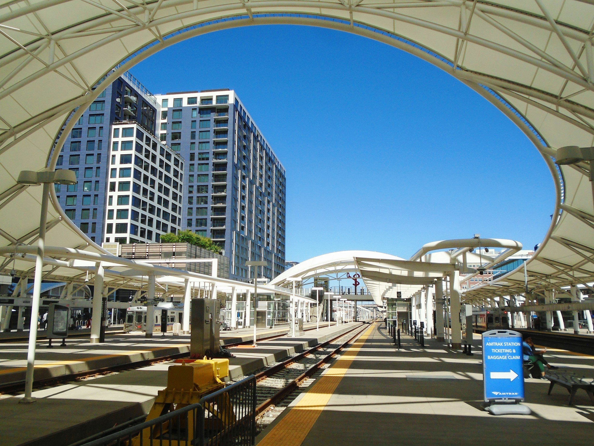 Union Station in Denver CO train station platform with tracks, ticketing, and baggage claim signs, surrounded by modern high-rise buildings under a clear blue sky.