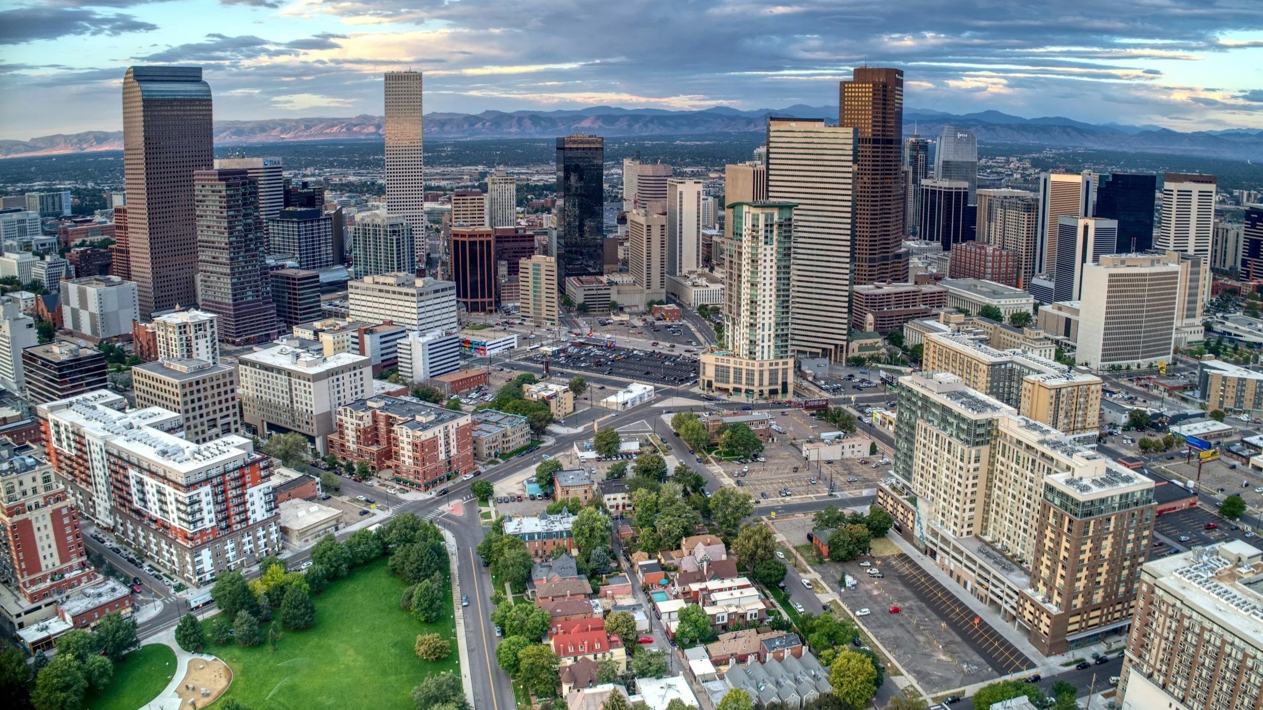 A city skyline with high-rise buildings, office towers, and urban streets seen from an aerial view, with mountains in the background and a partly cloudy sky overhead.