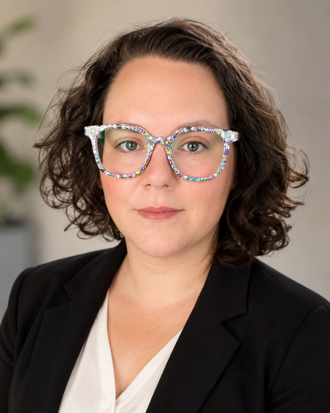 A woman with curly brown hair wearing colorful, patterned glasses and a black blazer.