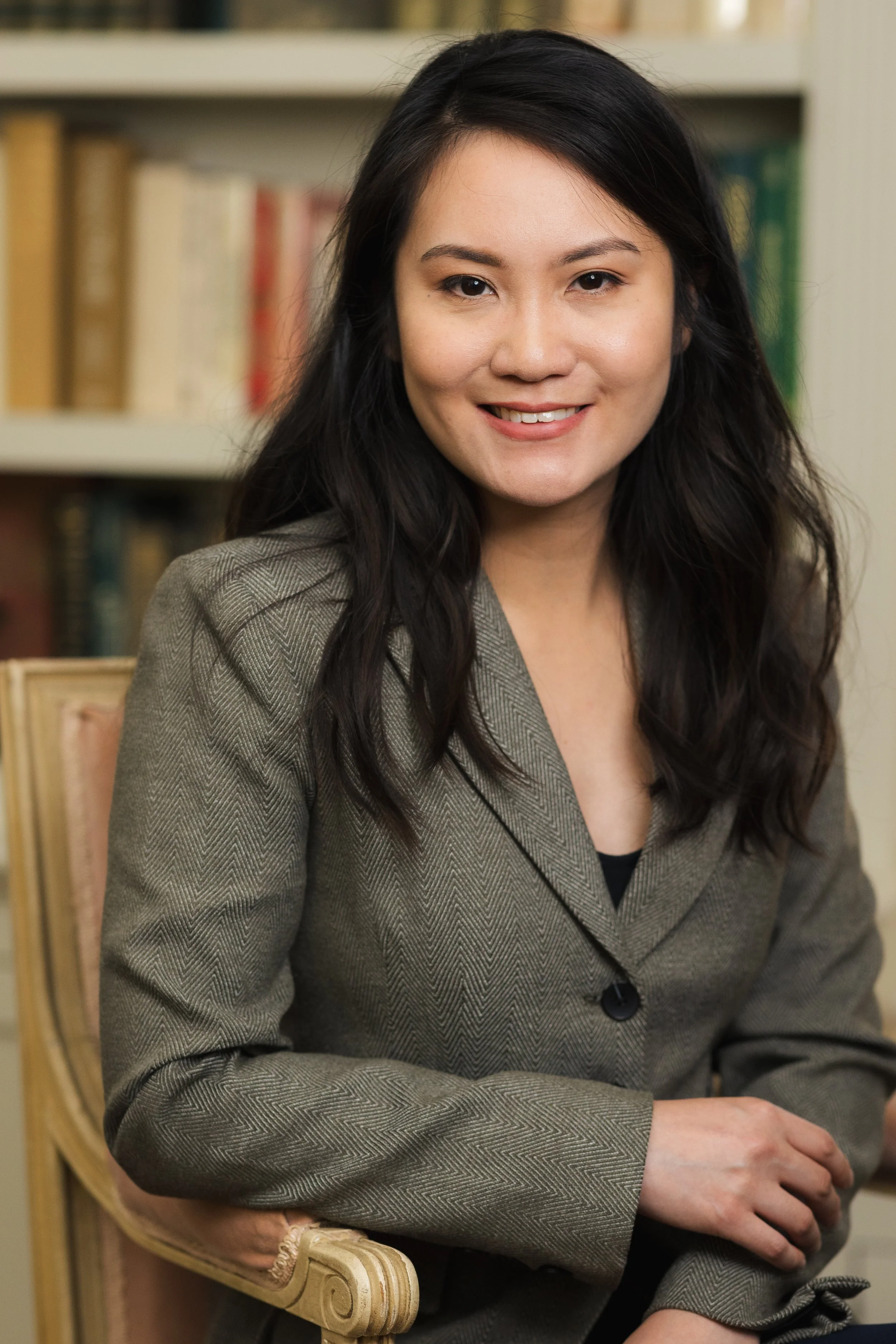 A woman with long dark hair sitting on a gold ornate chair, wearing a gray blazer, smiling in front of a bookshelf with books.