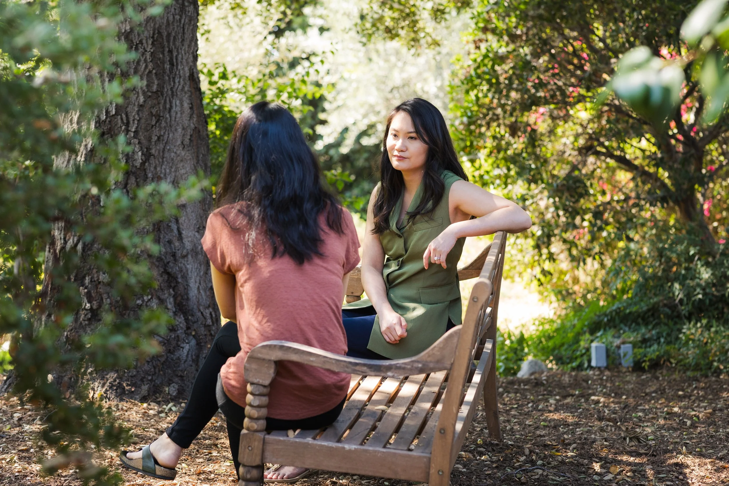 Two women are sitting on a wooden park bench, engaged in conversation, surrounded by greenery and trees, with sunlight filtering through the leaves.