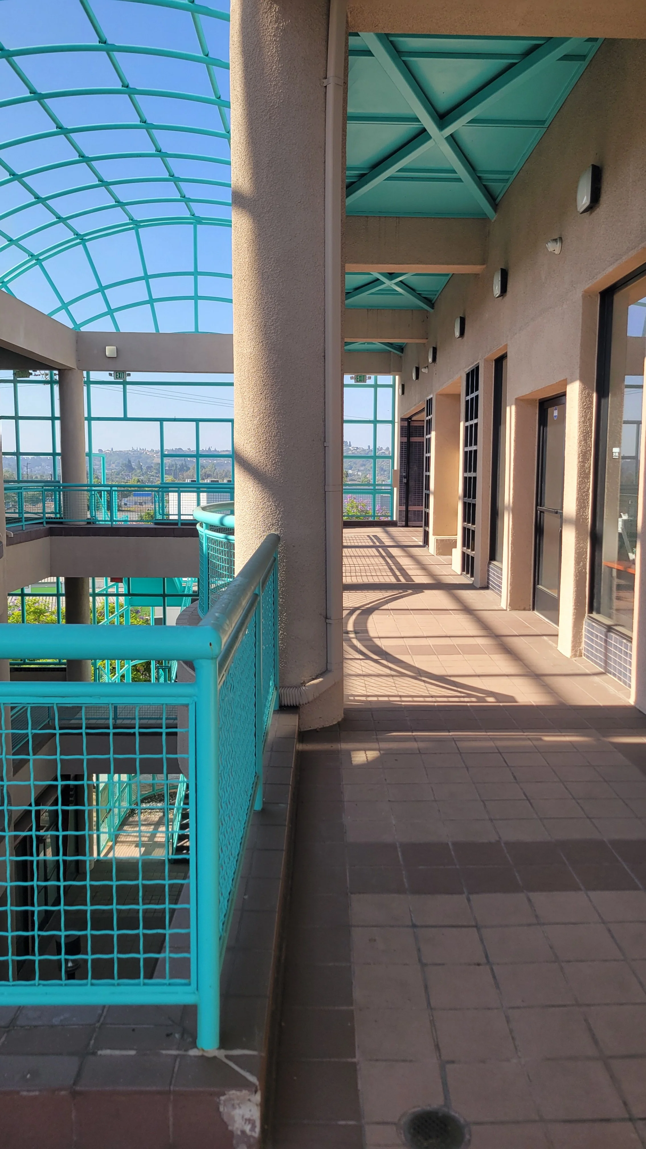 View of an outdoor balcony area with beige walls, aqua-colored metal roofs, and turquoise railing. Multiple doors are along the wall, with shadows cast on the tiled floor. The sky is blue, and cityscape can be seen in the background.