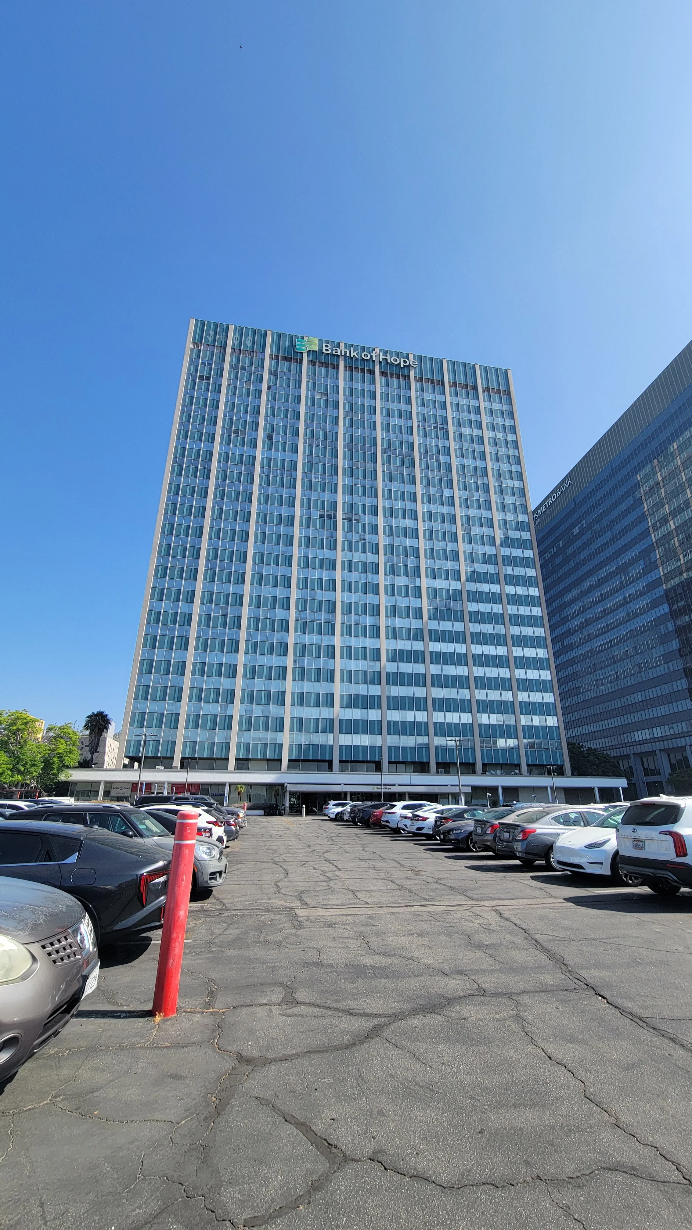 Tall office building with a sign that reads 'Bank of Hope,' a parking lot in the foreground filled with cars, and a clear blue sky.