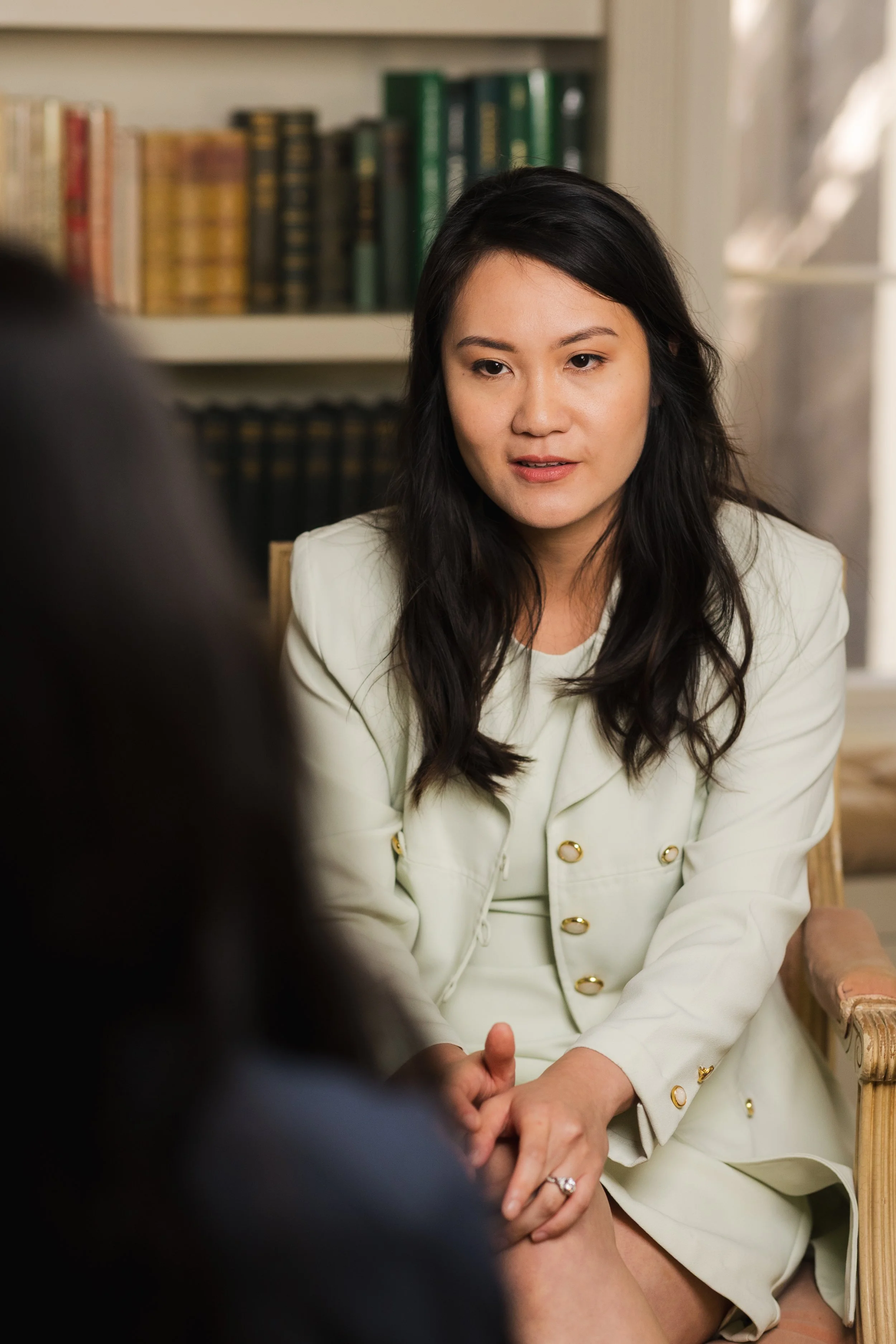 A woman in a white blazer with gold buttons appears to be engaged in a conversation, sitting in a room with a bookshelf filled with green and brown books in the background.