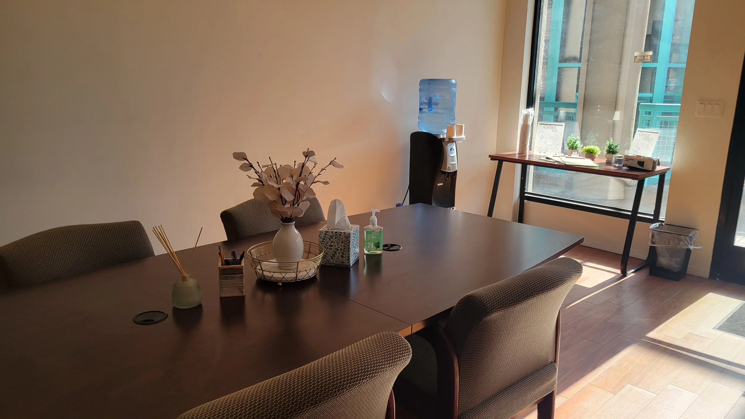 Meeting room with a large wooden table surrounded by chairs, a vase with artificial flowers, a bottle of hand sanitizer, tissues, and a small container with snacks. A water dispenser is near the window with sunlight.