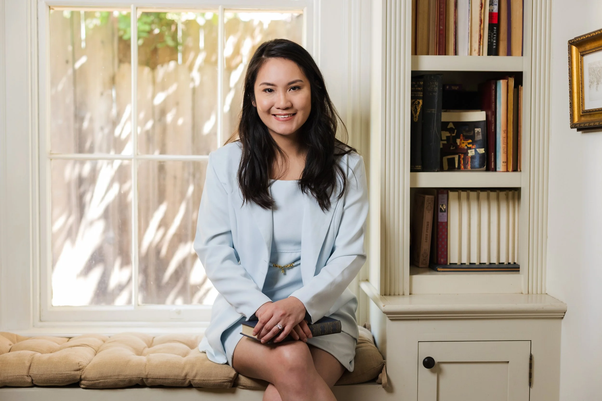 A woman with long dark hair, wearing a light-colored blazer and shorts, sitting on a cushioned window seat with a book in her lap, smiling at the camera in a cozy room with beige carpet, white walls, a bookshelf with various books, and a large window with sunlight coming in.