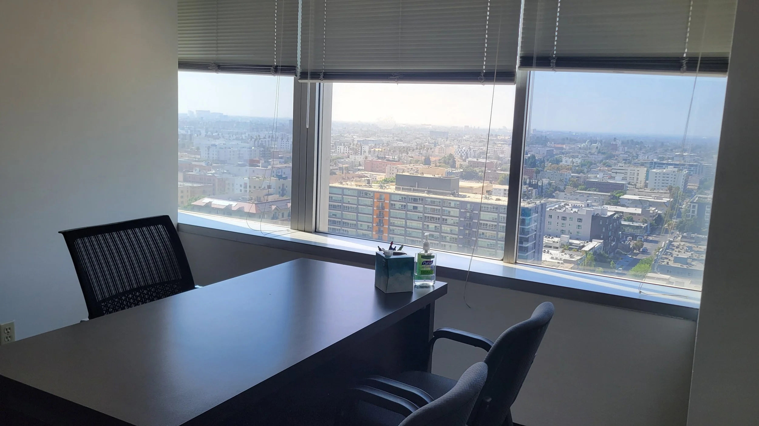 Office meeting room with a window view overlooking a city skyline, featuring a desk, a black office chair, and cleaning supplies on the desk.