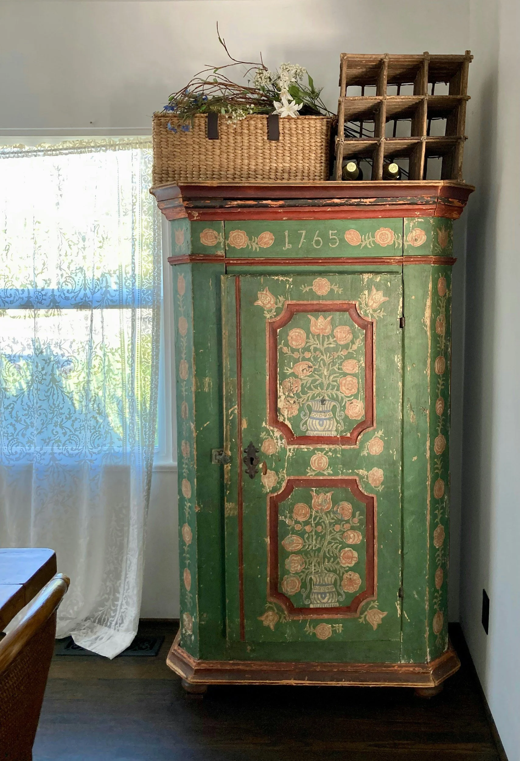 An antique green wooden cabinet with floral and urn painted designs and the year '1765' written above the door, situated in a room next to a window with sheer lace curtains. On top of the cabinet are a woven basket with white flowers and greenery, and a small wooden wine rack with three wine bottles.