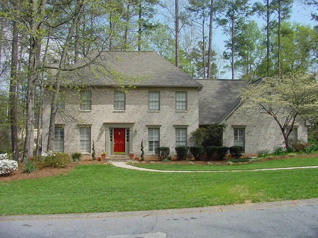 A two-story brick house with a red front door, surrounded by trees and a landscaped lawn.