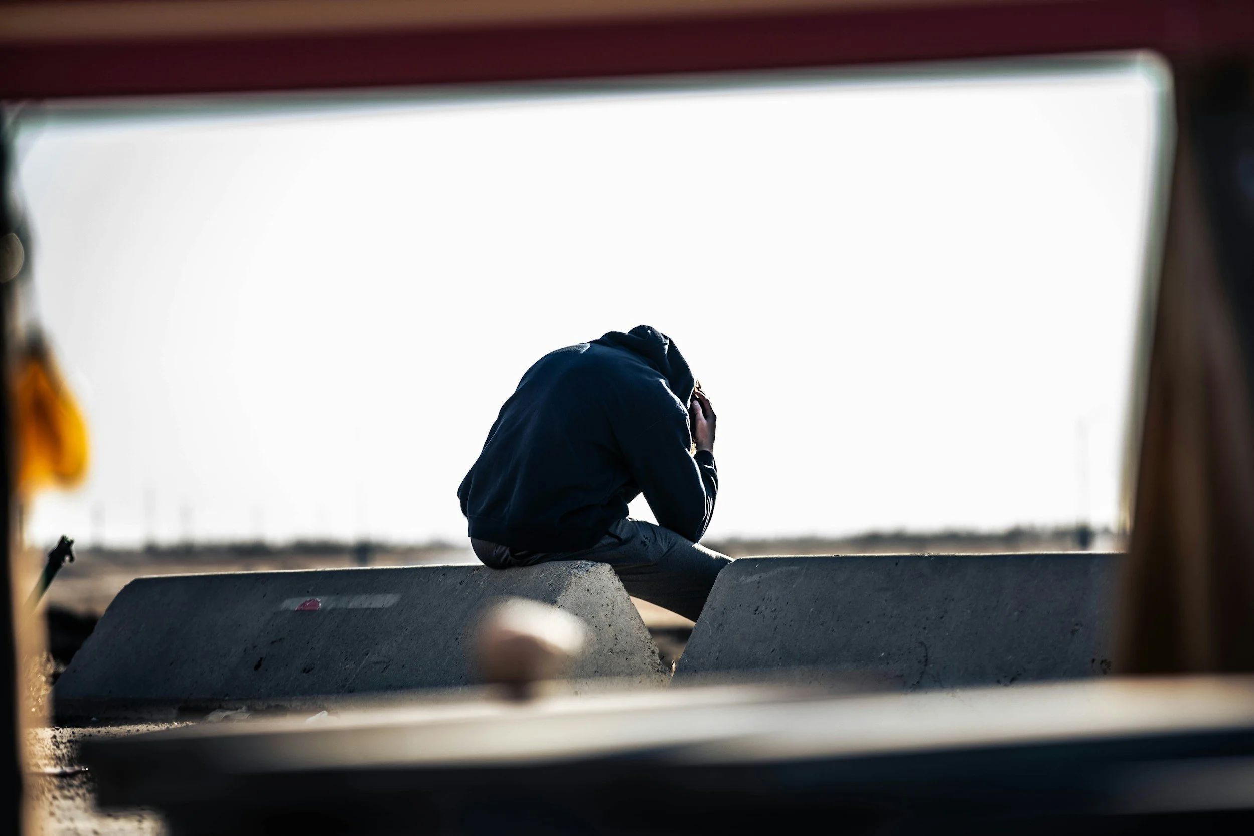 A person sitting on a concrete barrier near a road, with their head bowed and hand on their face, wearing a dark jacket, possibly consoled or contemplative, with a blurred background.