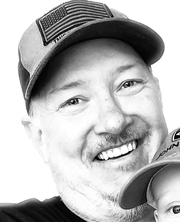 Black and white photo of a man smiling, wearing a baseball cap with an American flag patch on it, and part of another person's face is visible in the lower right corner.