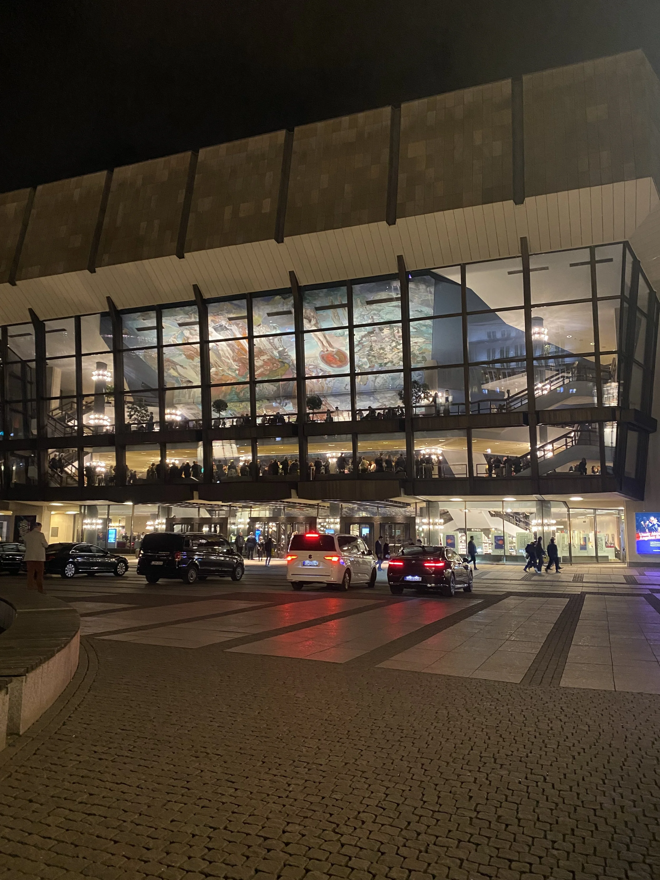 Ein mehrstöckiges Gebäude mit vielen Fenstern, durch die Einblick in ein beleuchtetes Innere mit zahlreichen Menschen und Kunstwerken möglich ist. Im Außenbereich sind Parkplätze mit Autos und Fußgänger sichtbar.