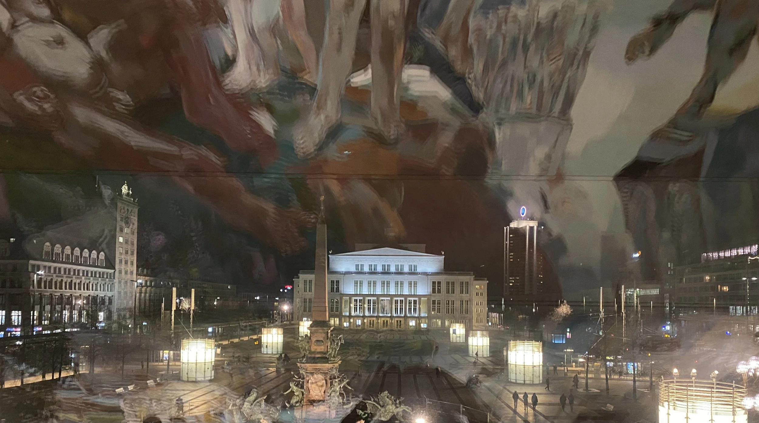 Stadtplatz bei Nacht mit beleuchteten Gebäuden, Skulptur im Vordergrund, reflektierende Oberflächen und städtischer Skyline im Hintergrund.