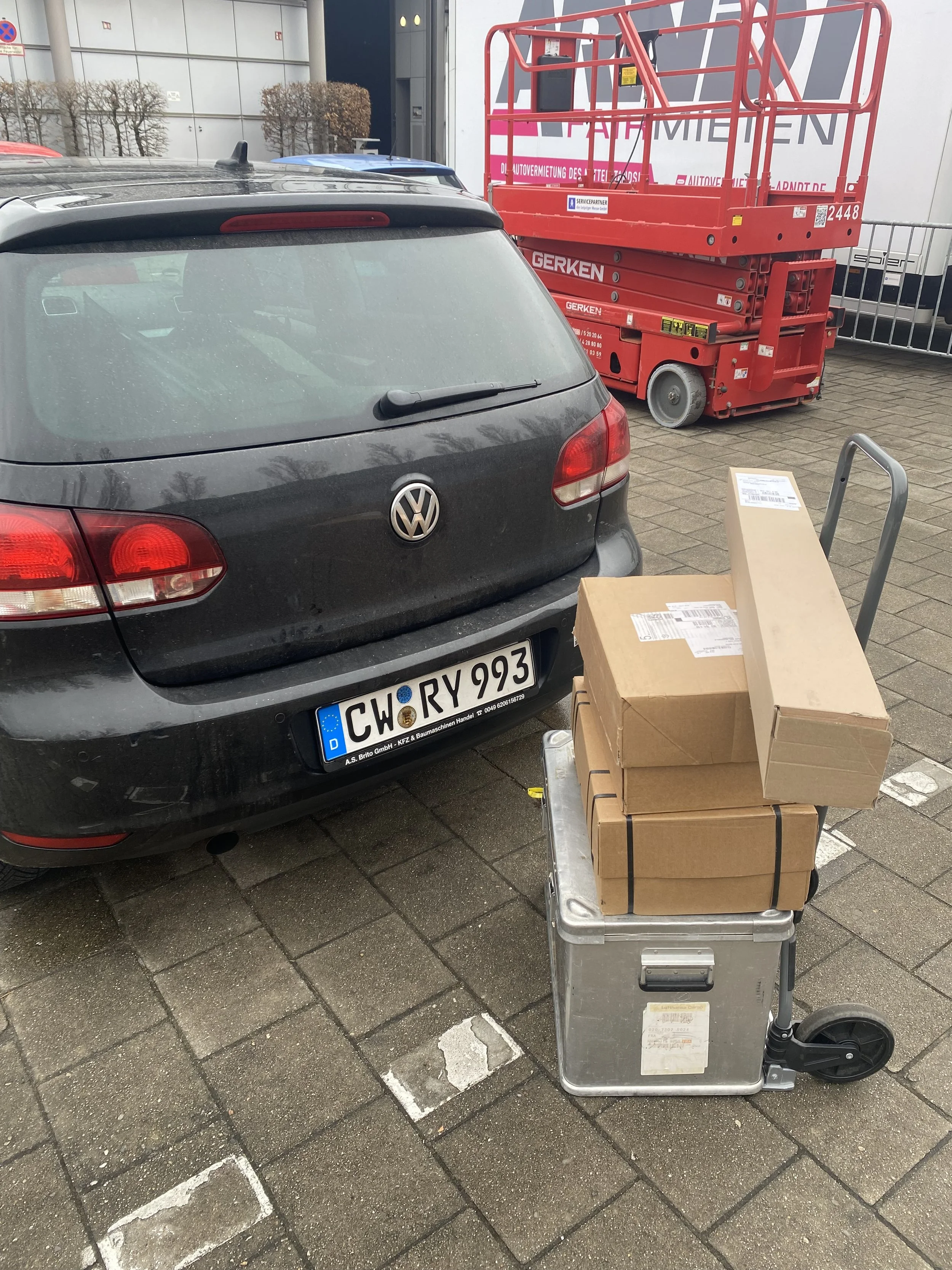 Ein schwarzer Volkswagen Polo mit deutschen Kennzeichen steht auf einem Parkplatz. Neben dem Auto ist ein Handwagen mit mehreren Kartons. Im Hintergrund sind eine rote Hebebühne und eine Gebäudefassade sichtbar.