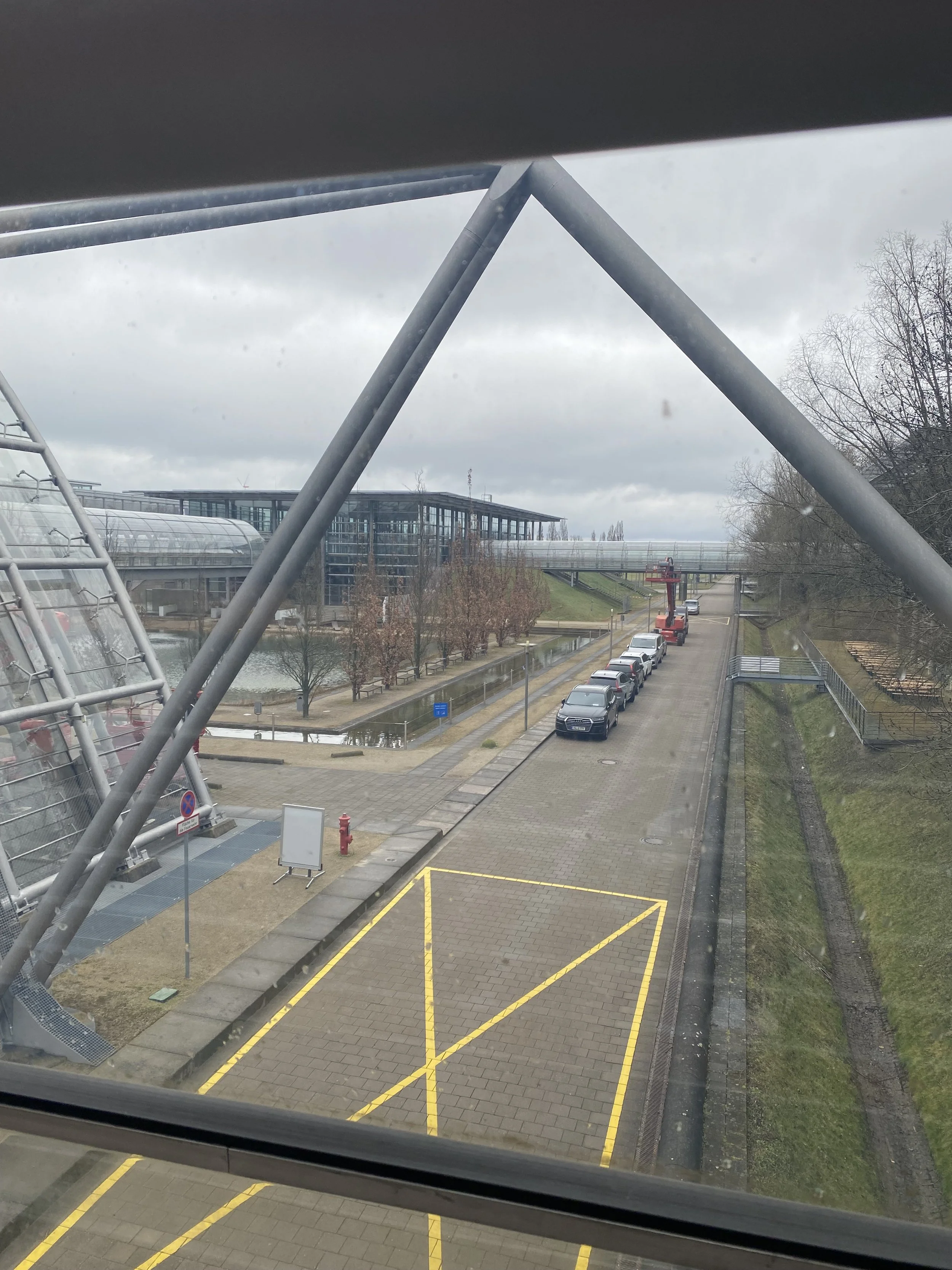 Blick durch ein Fenster auf einen Parkplatz mit gezeichneten Linien, einer Straße mit geparkten Autos, Bäume und eine moderne Gebäudefront mit großen Fenstern. Das Wetter ist bewölkt.
