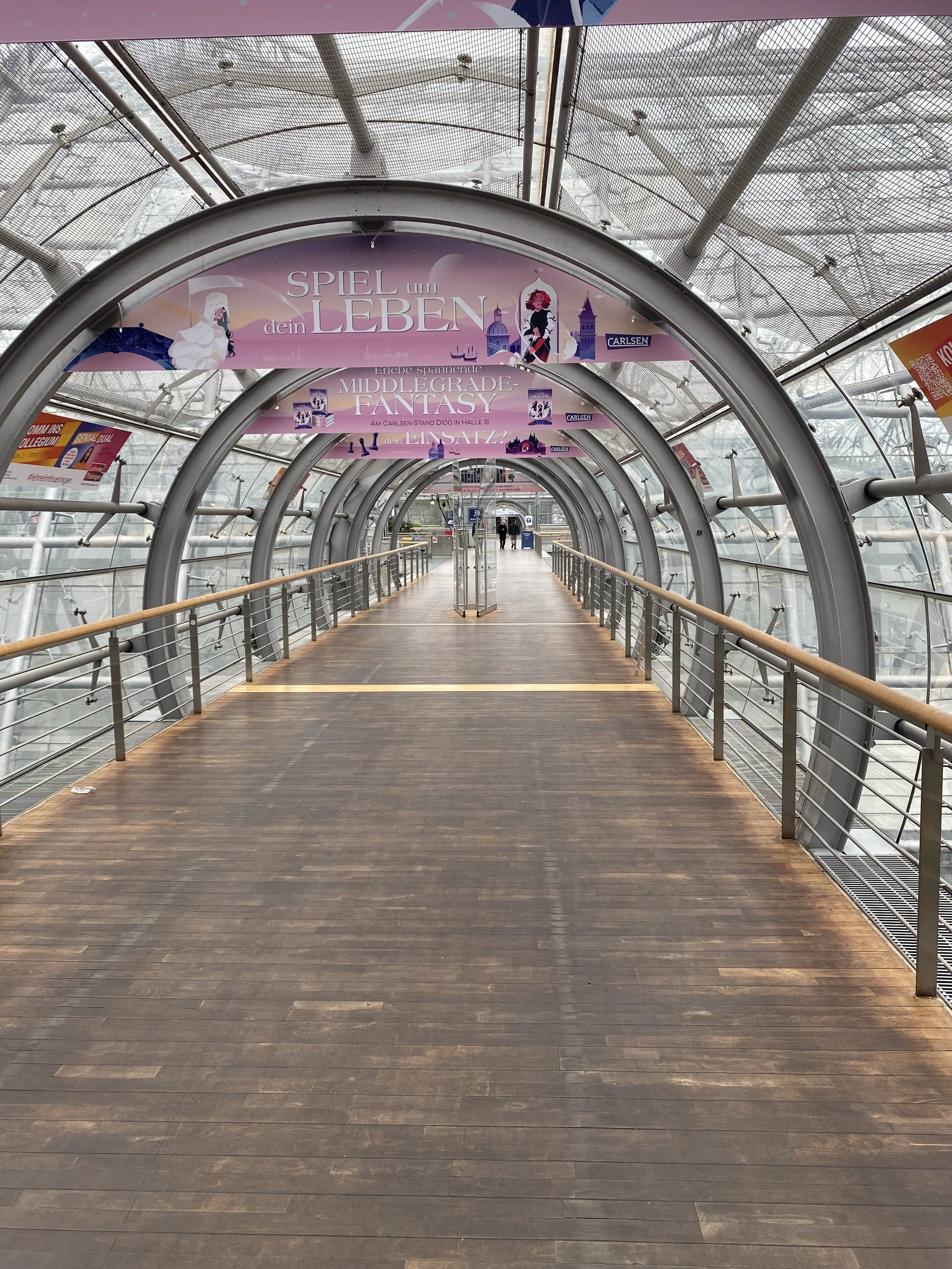 Innenansicht eines gläsernen Tunnels mit Holzfußboden, entfernt zwei Personen stehen vor einem Eingang, an der Decke hängen Banner mit deutschem Text, Gebäude im Hintergrund sichtbar.