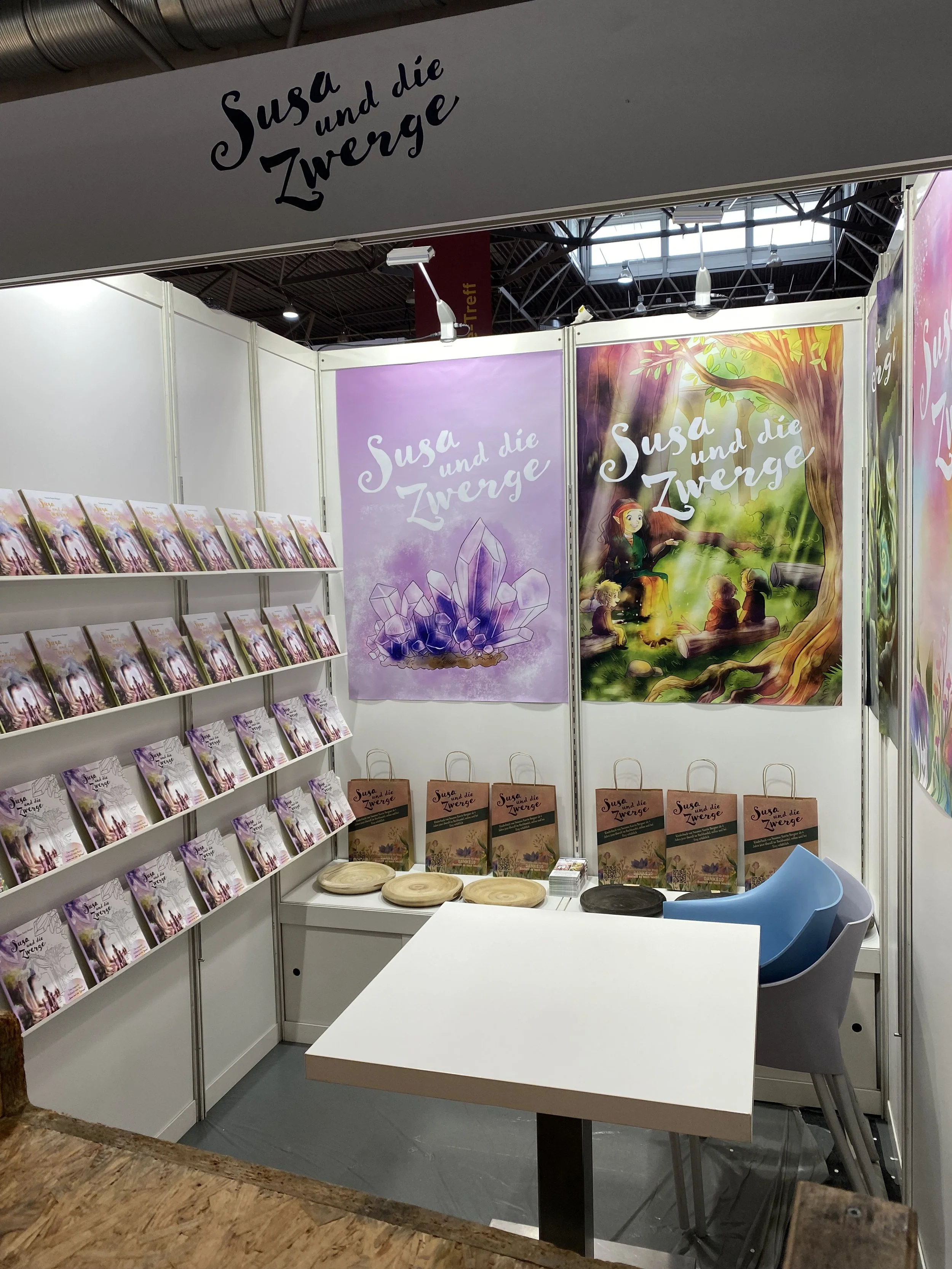 Ausstellungsstand mit Büchern und Postern der Kinderbuchreihe 'Susa und die Zwergen' bei einer Buchmesse. Das Logo ist auf den Postern und einem Wandbanner sichtbar, das eine Illustration einer freundlichen Waldszene mit einer kleinen Figur zeigt.